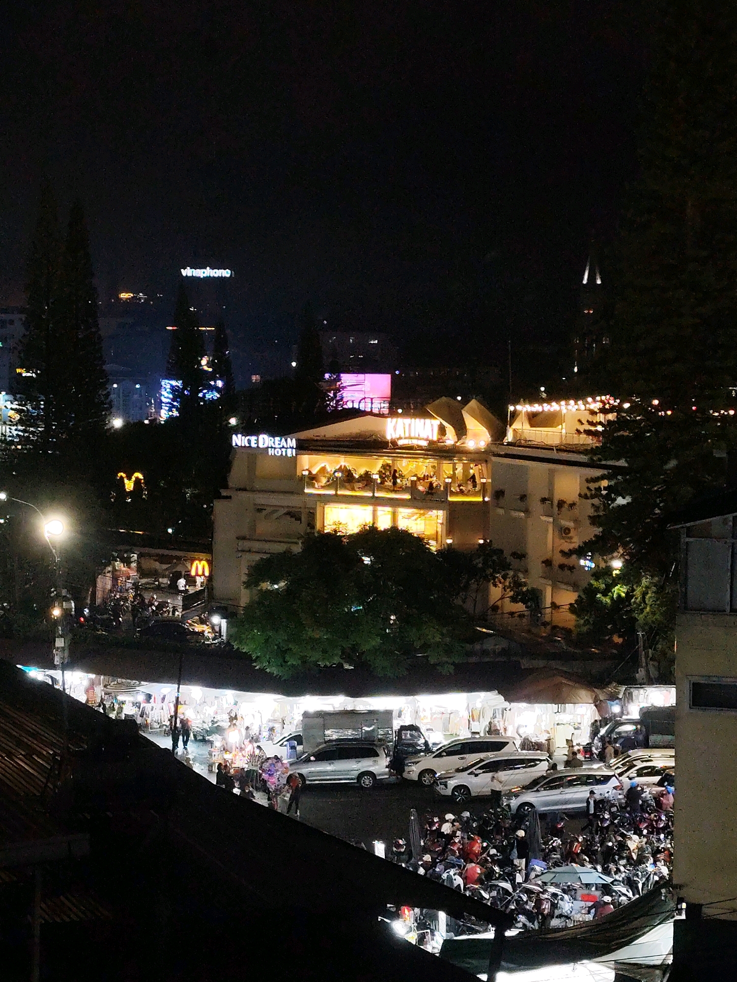 This is a night-time, high-angle shot of a busy street in Da Lat, Vietnam. The scene is illuminated by a mixture of artificial lights from buildings, streetlights, and illuminated signage. In the foreground, a dark, slanted roofline partially obscures the view.
Further into the image, a bustling street market is visible with numerous stalls and vendors under bright lights, selling various goods. People are seen walking and interacting within the market. Several vehicles, including cars and many motorbikes, are parked and in motion on the street.
Dominating the middle ground is a multi-story building with a prominent sign reading "NICE DREAM HOTEL". The building is lit from within, showcasing glowing windows and illuminated balconies with decorative string lights. Adjacent to the hotel, another building displays a sign that appears to read "KATINAT". Further in the distance, other buildings with illuminated windows and signs are visible, contributing to the urban nightscape. A bright McDonald's sign is also visible. Trees dot the landscape, with some illuminated and others silhouetted against the dark sky. The overall impression is one of vibrant urban nightlife.
