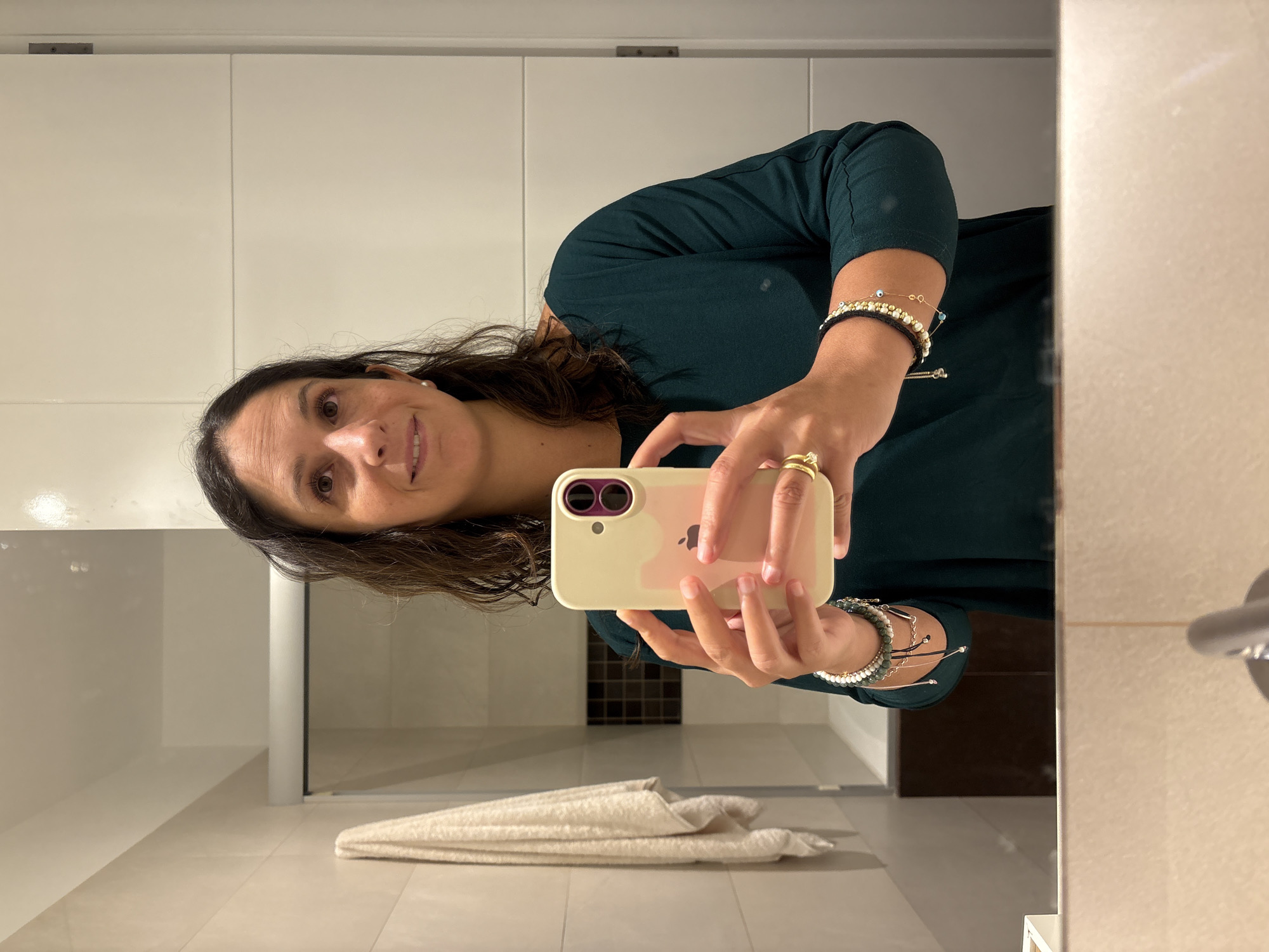 A woman with long, dark, wavy hair takes a mirror selfie in what appears to be a bathroom. She is wearing a dark green long-sleeved shirt and multiple bracelets on her left wrist, including beaded and metallic styles. She is holding a pale yellow phone with a pink insert in front of her, presumably to take the photo. Her right hand, adorned with a gold ring, is also visible. The background shows tiled walls, a towel hanging on a rack, and a glimpse of a bathtub. The mirror reflects the woman, the phone, and the surrounding environment.
