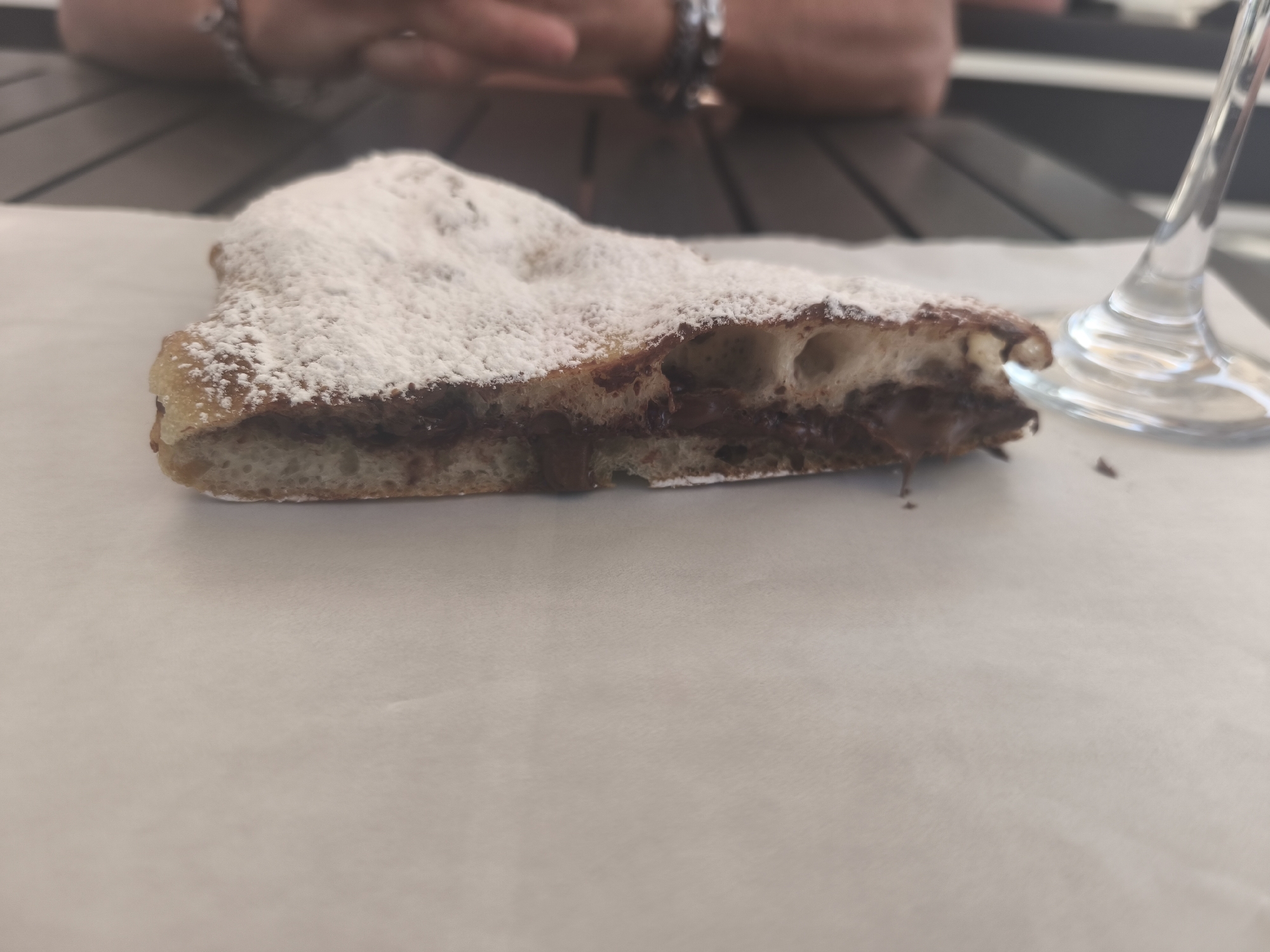 A close-up, slightly overhead shot shows a slice of dessert, possibly a calzone or pastry, filled with chocolate and dusted with powdered sugar. The dessert rests on a sheet of white paper. In the background, blurred figures of people are visible, along with a table and a clear glass. The scene appears to be outdoors, possibly at a cafe or restaurant in San Salvo, Italy. The lighting is soft and natural.
