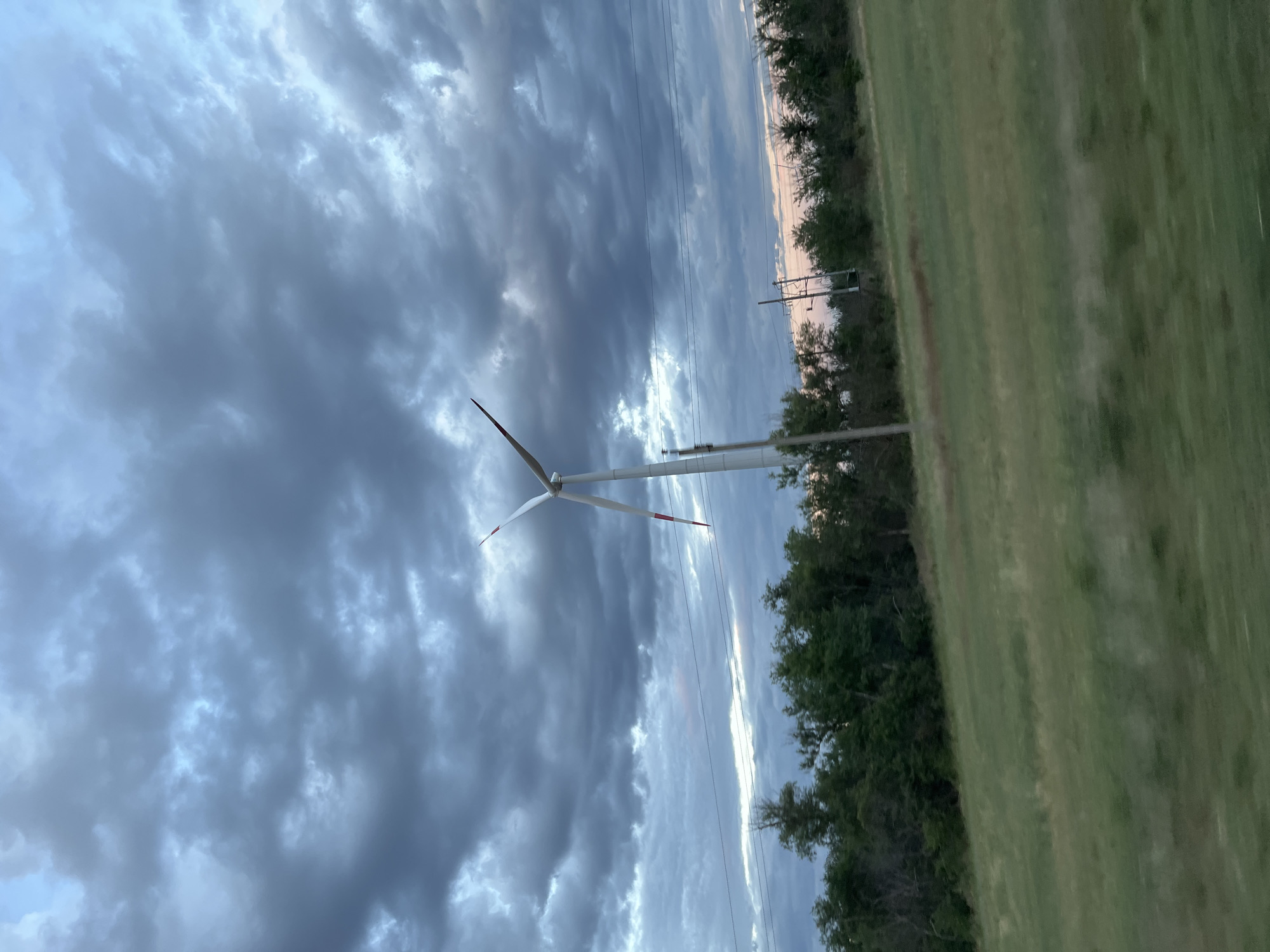 An outdoor, eye-level, medium shot captures a wind turbine under a dramatic, overcast sky, a classic view of Zimovniki, Russia. The turbine, white with red accents at the tips of its blades, stands tall in the center of the frame. Its three blades are partially obscured by the cloudy sky. A field of green grass, slightly blurred by motion, fills the foreground. In the middle ground, a line of dense green trees runs horizontally across the image, with power lines and poles visible above them. The sky is dominated by dark, textured clouds, suggesting a potentially stormy or twilight atmosphere. The overall impression is one of vast, open landscape and a powerful, modern energy source against a natural backdrop.