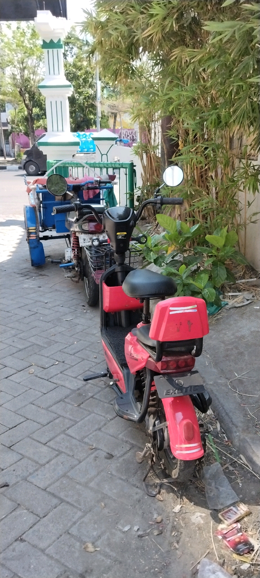 The image depicts a row of parked electric scooters on a paved sidewalk in Surabaya, Indonesia. The foreground features a red electric scooter with a black seat, and a smaller black seat behind it. The scooter has a license plate that reads "EXOTIC". Behind it, another scooter, possibly also electric, is attached to a blue trailer. This scooter has a black handlebar, a black seat, and a red and blue colored cargo area in the trailer. Further down the row, a black three-wheeled vehicle with a blue cargo box is partially visible. In the background, a white and green striped monument or structure stands near some lush green bamboo trees and other foliage. The ground is paved with gray brick-like stones, and there are some scattered leaves and debris on the sidewalk and adjacent dirt area. The scene suggests a typical urban environment in Surabaya, Indonesia, with a focus on personal transportation and possibly light cargo transport.