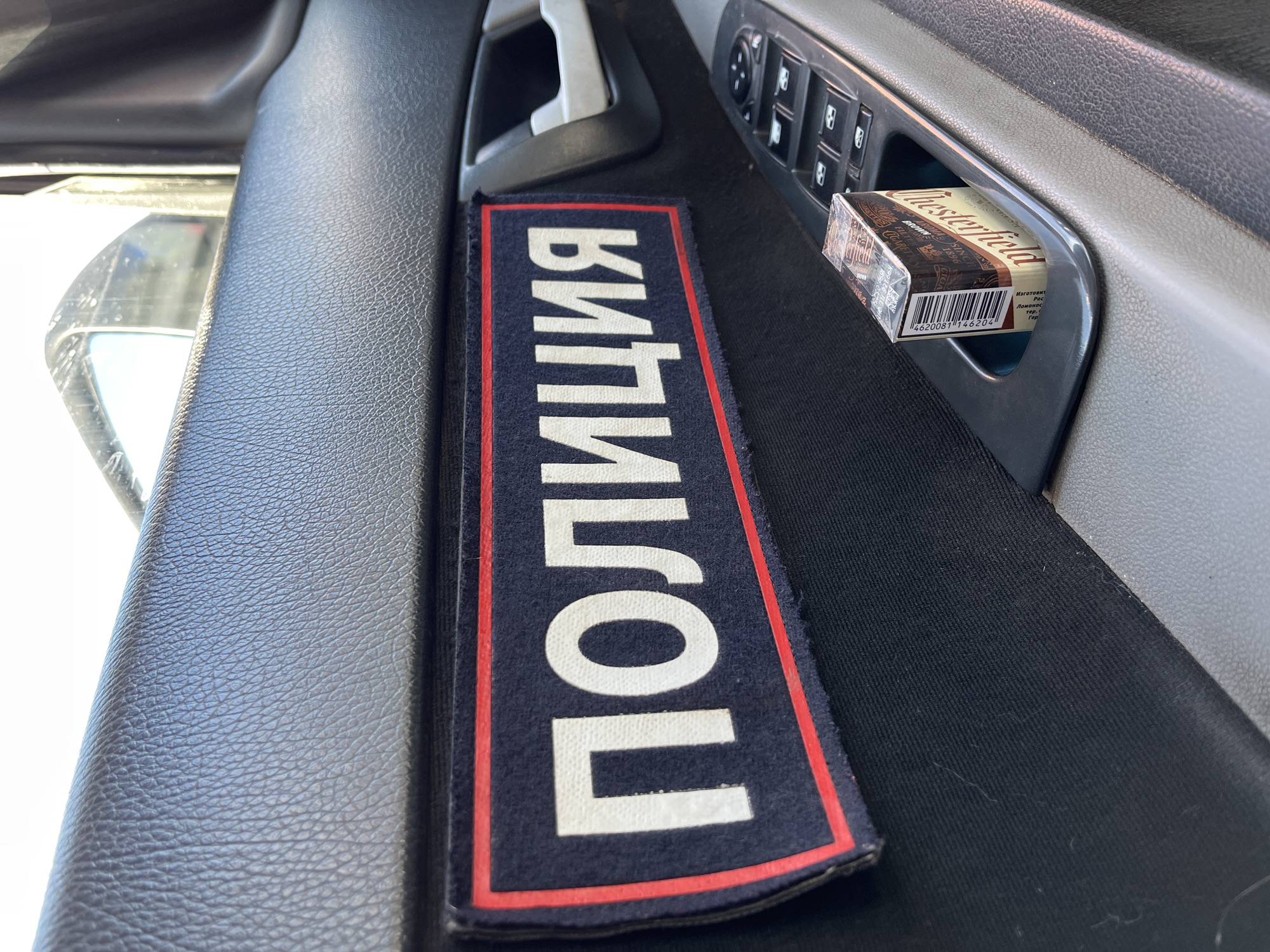 The image depicts the interior of a vehicle, specifically the passenger side door panel, in Chaikovsky, Russia. A rectangular dark blue patch with white Cyrillic text "ПОЛИЦИЯ" (POLITSIYA, meaning "POLICE") and a red border is affixed to the black fabric insert of the door. Below this patch, a pack of Chesterfield Brown cigarettes, still sealed in plastic, rests vertically in a molded compartment on the dark gray plastic door panel. Adjacent to the cigarette pack is a control module with multiple black buttons, likely for power windows. A light gray door handle is visible further back. The top portion of the door panel is textured dark gray plastic. A reflection in the exterior side mirror, visible through the partially open window, indicates an outdoor, daytime setting. No individuals are visible.