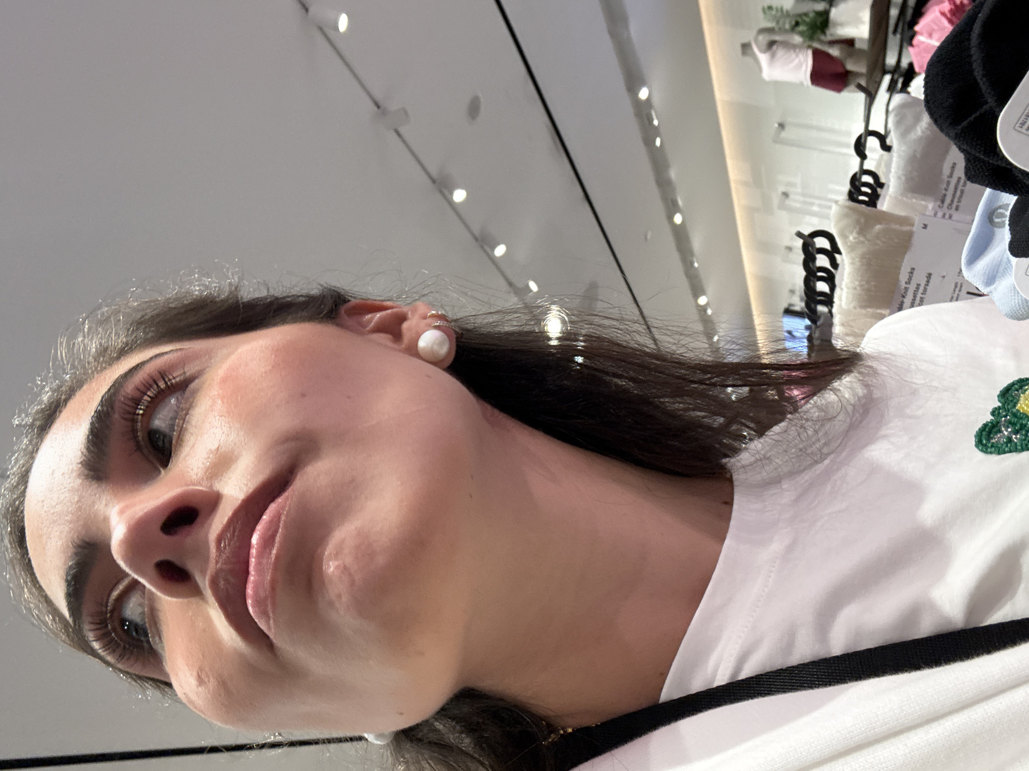 A woman with dark hair and fair skin is looking up towards the camera. She is wearing a white top with a black stripe, and pearl earrings. In the background, socks and clothing on hangers are visible, suggesting a retail environment. The setting is likely a store located in San Antonio, United States of America, but this detail is inferred from the prompt rather than directly visible in the image.