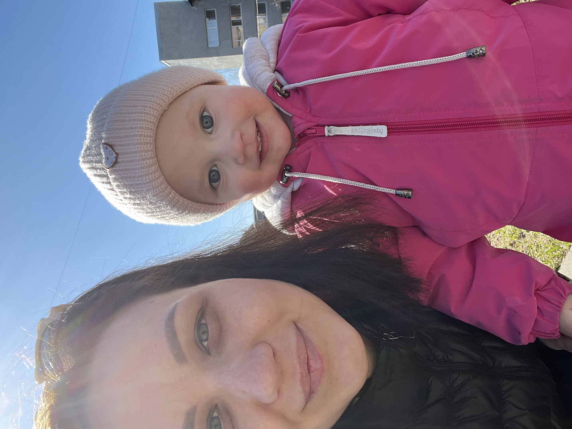 A selfie taken at an angle upward shows a woman and a baby. The woman has dark hair and fair skin, wearing a black jacket. The baby, wearing a light-colored hat and pink jacket, is positioned above the woman, partially obscuring the view. The sky is visible in the background, along with a building. Based on architectural style and geographic location, it is possible that this photo was taken in Perm, Russia.