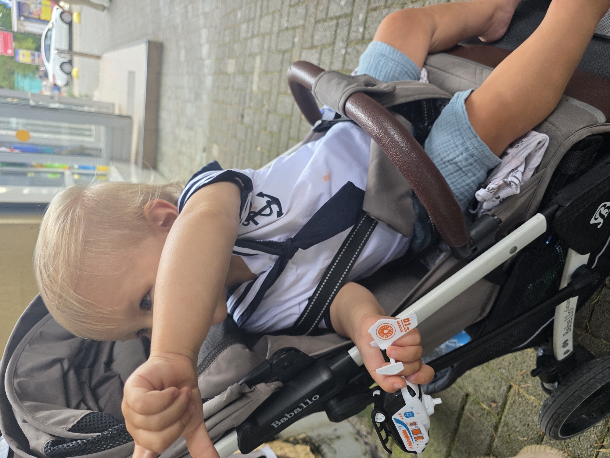 A blond-haired toddler wearing a white and navy sailor shirt and light blue shorts is sitting in a stroller. The toddler is holding a small toy helicopter in their right hand and pointing outwards with their left arm. The stroller is a dark gray color with a brown handle. The background shows a cobblestone pathway and a building with glass doors, suggesting an outdoor urban environment, possibly near a shop. The weather appears overcast. The image is taken from a slightly high angle, focusing on the child and the stroller.