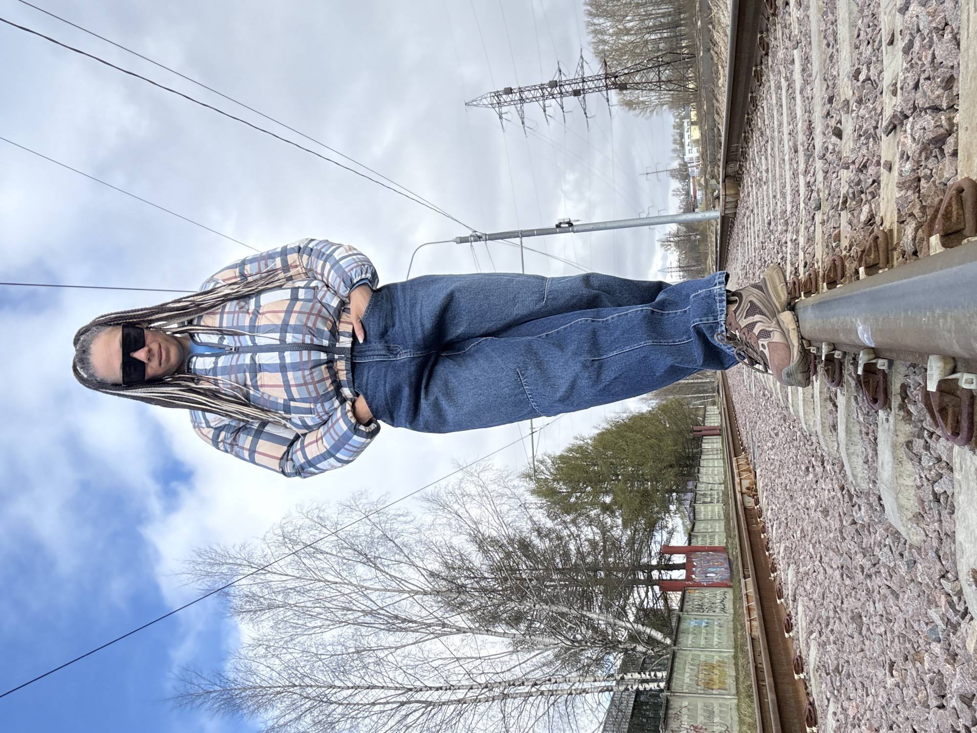A person with braided hair, wearing sunglasses, a plaid jacket, and jeans, stands on a train track. The track is surrounded by gravel. Behind the person is a sky with clouds and trees. A power line pole is visible in the background. A building with graffiti is to the left of the train track. This image appears to be taken in Rusanovo, Russia.