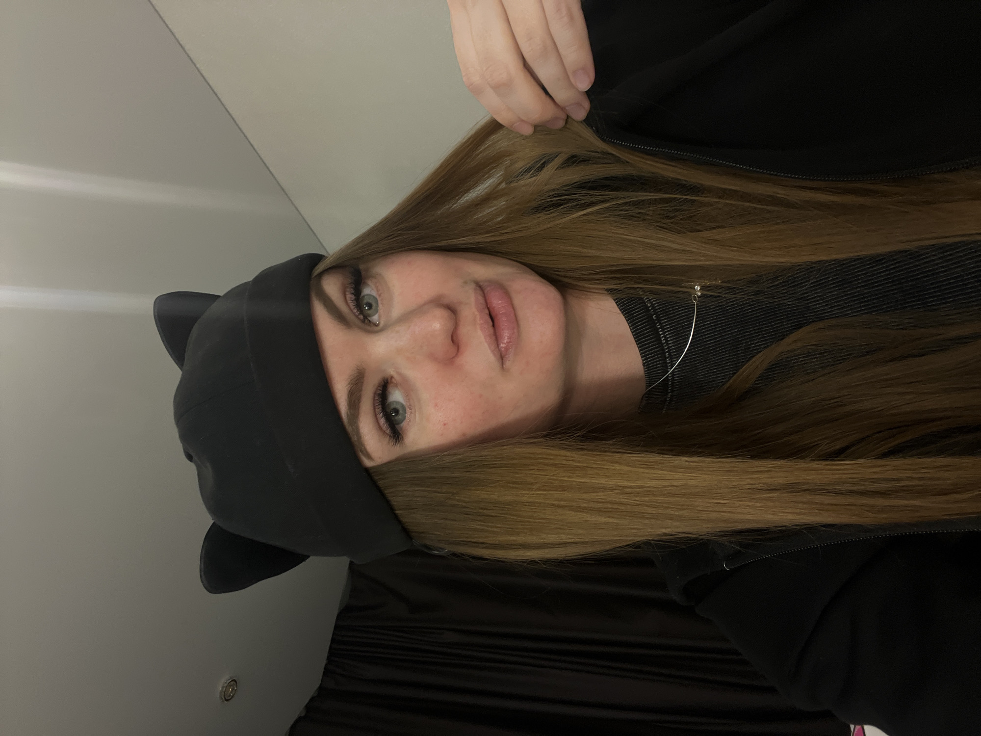 A woman with long, light brown hair and green eyes is wearing a black beanie with cat ears. She is also wearing a black hooded jacket over a dark gray striped shirt. Her face is partially covered by her hair and the beanie. The background consists of a light gray wall and a black curtain. The location is unknown.