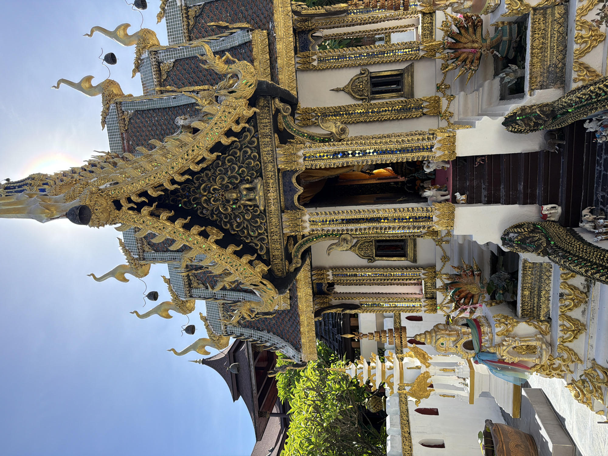 This is a low-angle, eye-level shot of a highly ornate, miniature Buddhist temple structure bathed in bright sunlight. The temple is adorned with abundant gold detailing, intricate carvings, and colorful mosaic tiles. The roof features multiple tiers, each decorated with golden mythical creatures and bells. A golden Buddha statue is visible within the temple's entrance. The structure is surrounded by a low white wall with golden decorative elements. To the left, a potted plant is visible. In the background, lush green trees and another, larger temple structure with a dark, curved roof can be seen against a clear blue sky. The overall impression is one of meticulous craftsmanship and vibrant religious symbolism.
