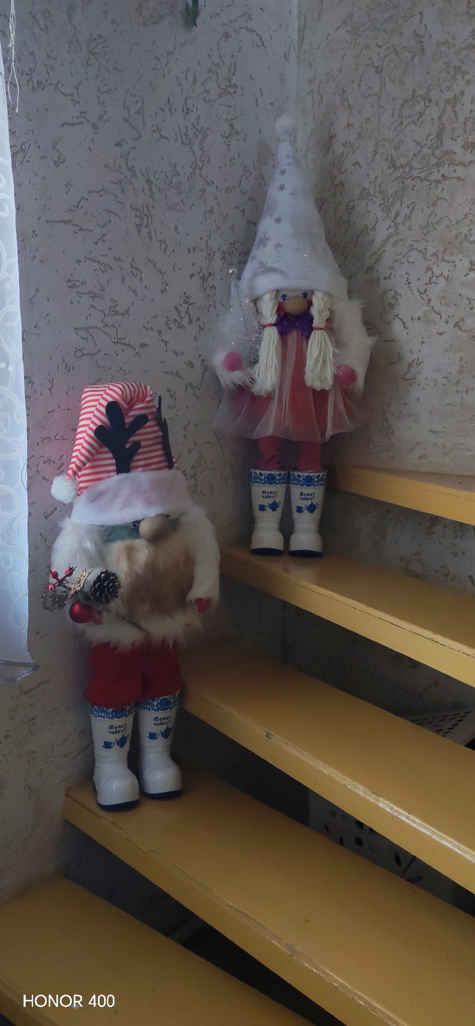 The image shows two Christmas gnome dolls positioned on wooden stairs. The gnome on the left wears a striped hat with reindeer antlers, a white beard, and red shorts, holding a decorative pine cone and berries. The gnome on the right wears a white star-patterned hat, a red dress with a tulle overlay, and has blonde braided hair. Both dolls wear white boots with blue floral patterns and Russian text. The background includes a textured white wall and a window with a white curtain on the left. The "HONOR 400" watermark is visible in the bottom left corner. The location is Noviy, Russia.