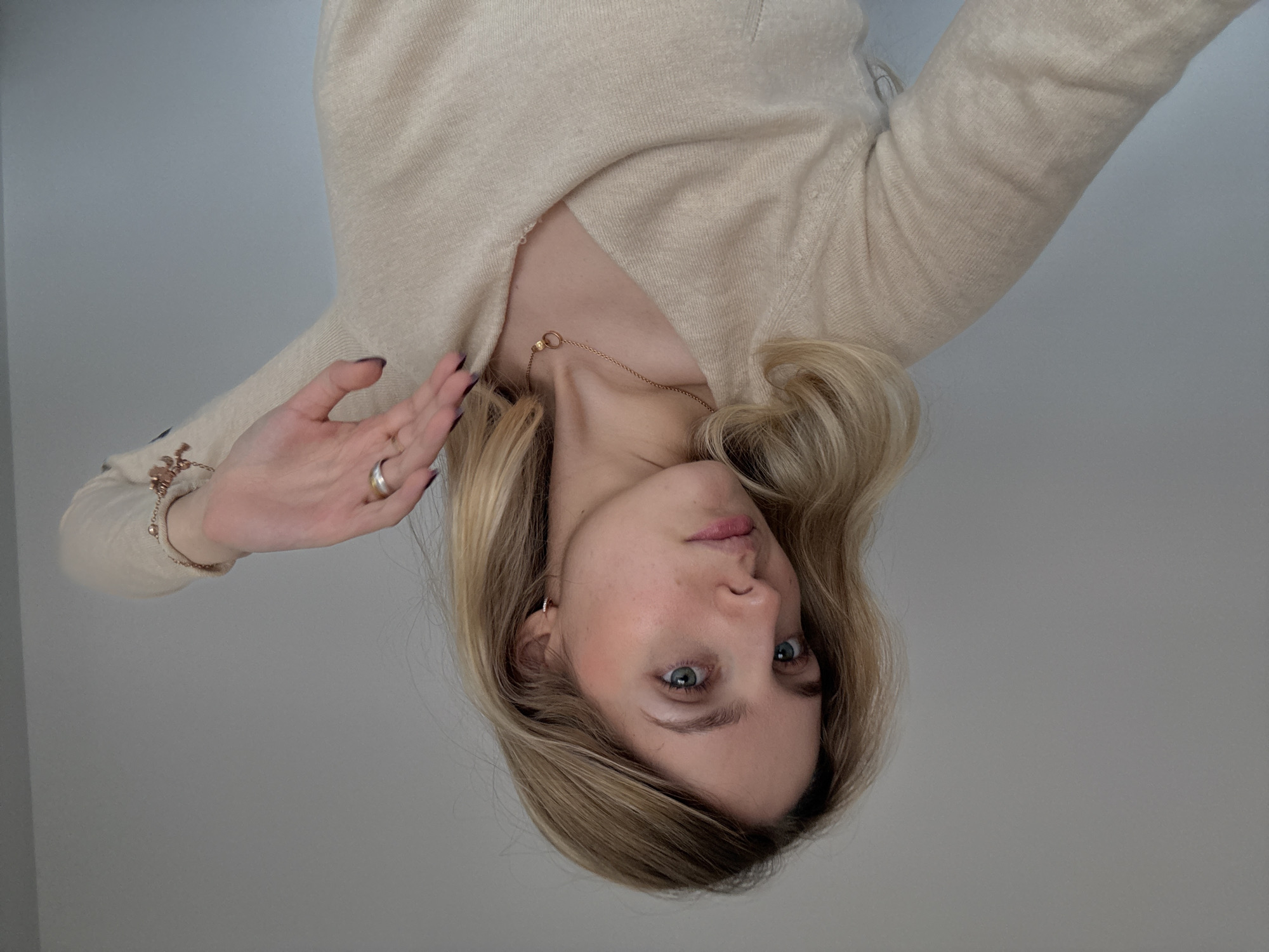 A woman with blonde hair and blue eyes is wearing a light-colored sweater and a necklace. She is looking directly at the viewer with a slight expression. Her right hand is raised with a ring on her ring finger, and a bracelet on her wrist. The background is a plain grey wall. The image is inverted. There is no information to identify the image location as Mexico City, Mexico.