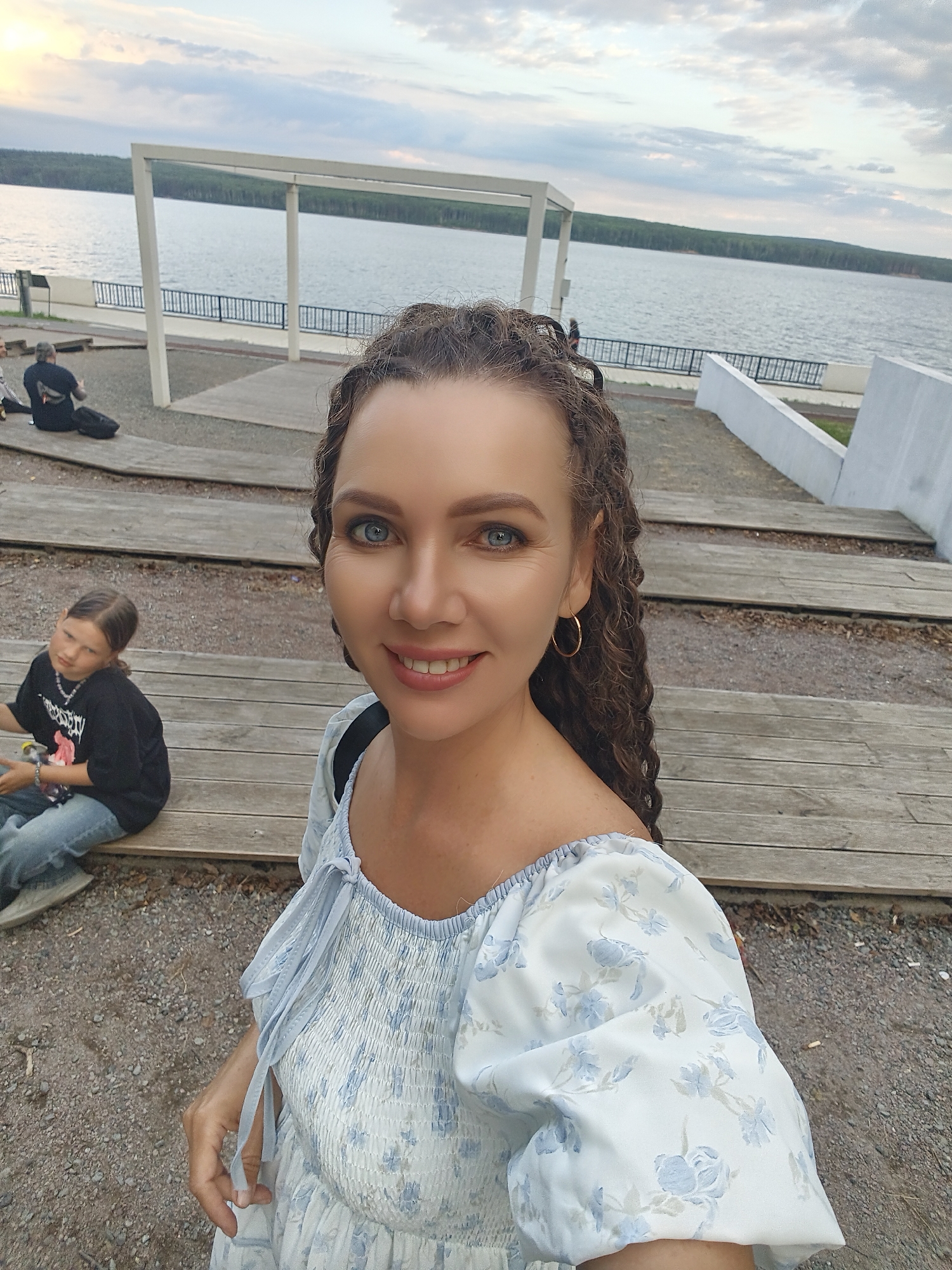 The photograph captures a woman taking a selfie in Chaikovsky, Russia, with a body of water and a treeline in the background. She is positioned in the foreground, smiling and looking towards the camera. She has curly dark hair and is wearing a white dress with a blue floral pattern and a light blue smocked bodice. A golden hoop earring is visible on her right ear. In the mid-ground, to her left, a young girl is seated on wooden bleachers, wearing a black t-shirt and blue jeans. Behind the girl, another person is visible sitting on the bleachers. The environment consists of wooden planks, gravel, and a paved area leading to the water. The sky is overcast with clouds.