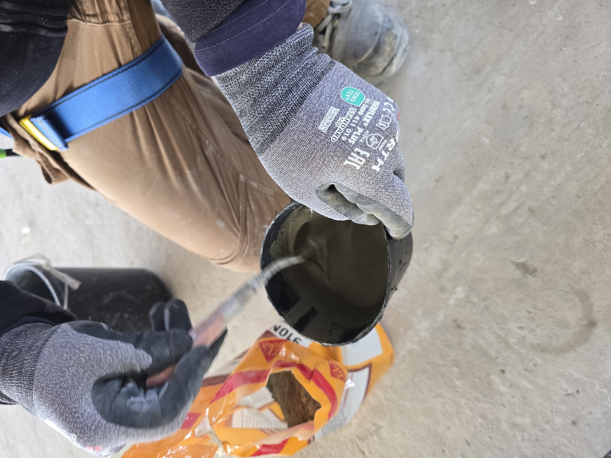 The image shows a construction worker mixing cement. The worker is wearing gloves and a safety harness. A cup filled with cement is positioned over a bag of cement mix. A tool is being used to mix the cement within the cup. The scene is located at an unspecified location.