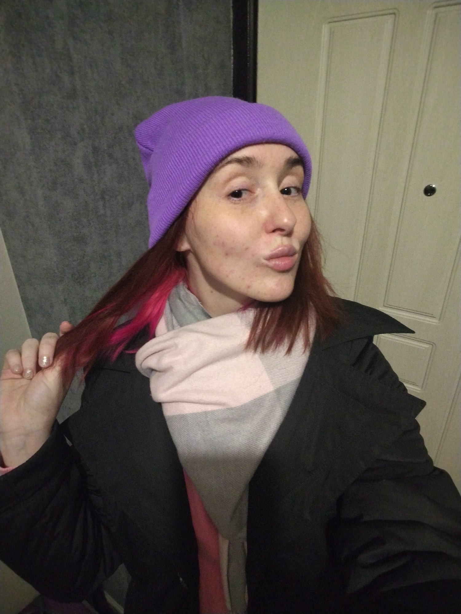 A woman is captured in a selfie-style shot, seen from the chest up, in an indoor setting. She has fair skin with visible blemishes or freckles and is making a playful puckered-lip expression. Her eyes are open and directed towards the camera.
She is wearing a bright, solid purple knitted beanie. Her hair, which is a reddish-brown color with distinct pink streaks, is visible below the beanie, and she is holding a lock of it with her right hand. Her fingernails have a light, possibly glittery, polish.
For clothing, she wears a black jacket or coat with a prominent collar. A thick, soft scarf in a light pink and grey plaid or checked pattern is wrapped around her neck, suggesting cooler weather.
The background is split between a dark grey, textured wall on the left and a light-colored, possibly off-white, paneled door on the right. A dark circular object, likely a peephole or door knob, is visible on the door. The lighting is artificial and indoors. There is no visible text in the image.