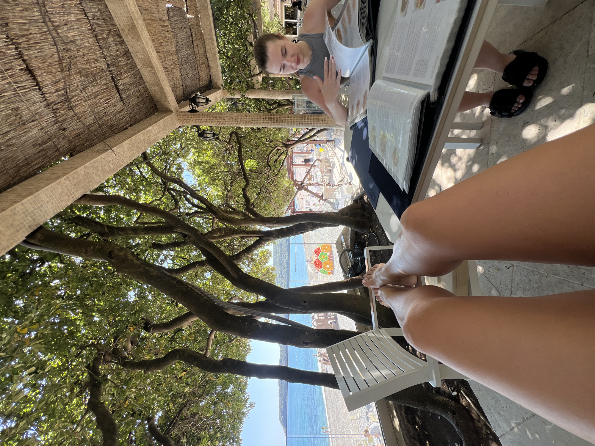 A woman is sitting at a table at a restaurant, looking at a menu. Another person's legs are extended towards the camera. The restaurant is under a canopy structure and a large tree, with a view of the beach and ocean in Prijevor, Montenegro.