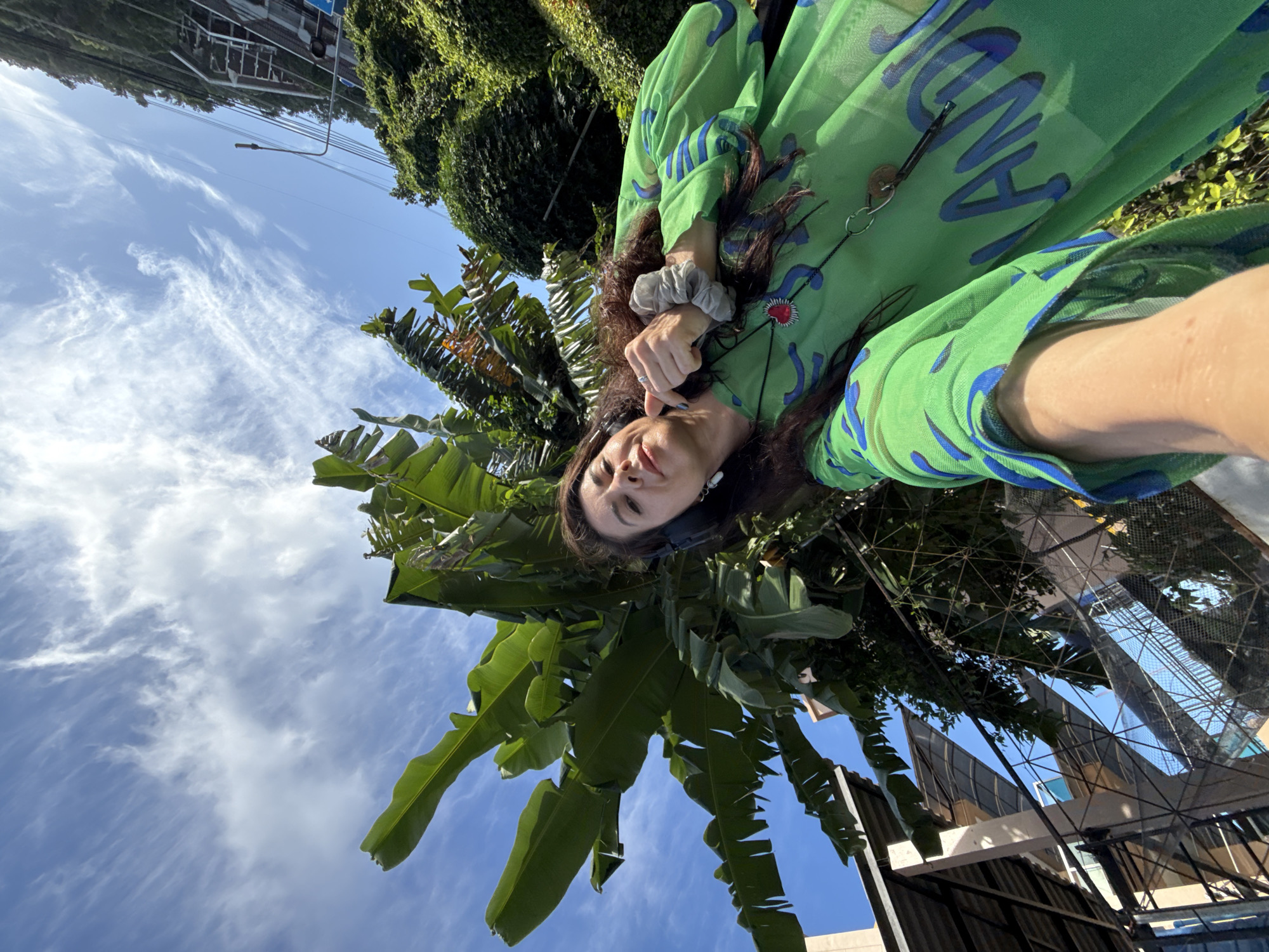 In Karon, Thailand, a woman with long, dark hair is taking a selfie outdoors. She is wearing a bright green, sheer top with blue abstract patterns and a heart-shaped pendant necklace. The woman is also wearing headphones and a scrunchie on her wrist. She is looking at the camera and smiling, with her right thumb up.
The background of the image is dominated by lush green foliage, including large banana leaves, and a clear blue sky with wispy white clouds. In the upper left corner, there is a glimpse of a building with a metallic roof structure, possibly a part of a resort or complex. Power lines and a street light are visible further in the background, indicating an urban or semi-urban setting. The perspective of the photo is from a low angle, looking upwards.