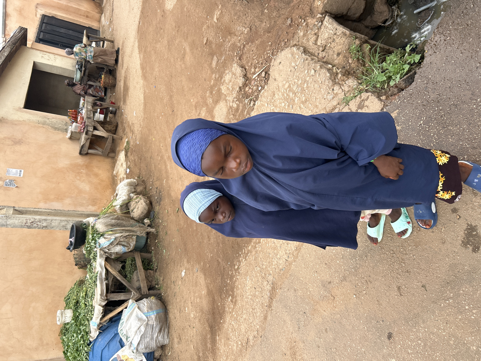 The image captures an outdoor street scene, likely in Jos, Nigeria, from an elevated perspective looking down. In the foreground are two young girls, possibly sisters, dressed in dark blue, flowing abayas or similar modest garments and hijabs. The girl in front wears a dark blue patterned under-cap, while the one behind her has a lighter blue, ribbed under-cap. Both girls look towards the viewer with neutral expressions. The girl in front wears dark blue slip-on sandals, and the girl behind wears light blue slip-on sandals, with red nail polish visible on their toes. A dark fabric with yellow embroidery is also visible near the front girl's feet.
The ground is a mix of packed dirt and worn asphalt or concrete. To the right, an open drainage ditch with rough concrete edges contains murky water and some green vegetation.
In the mid-ground and background, a vibrant market scene unfolds along a street. On the left, multiple wooden stalls display goods. One stall is heavily laden with large piles of fresh green leafy vegetables, alongside a black basin and various bundled sacks. Further back, two older women are seated at another stall. One woman in a patterned top and light brown skirt sits behind a counter displaying bottles and baskets. A light brown building with a dark, possibly metal, gate serves as the backdrop to the market activity. The scene appears to take place during daytime, under bright but soft light, suggesting an overcast day or morning/late afternoon, as there are no harsh shadows. Faint, unreadable text on a paper is visible on the building wall, and the word "Spard" can be seen on one of the light blue sandals.