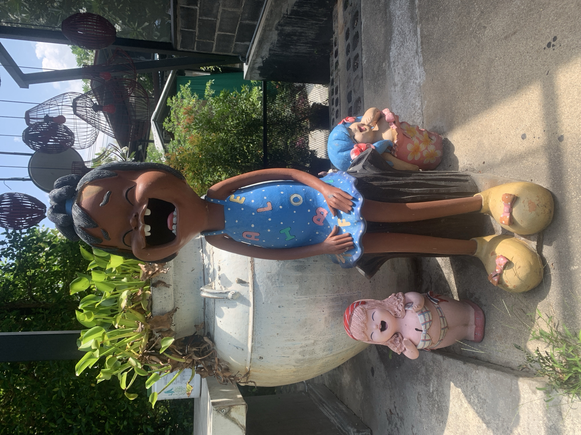 The image shows a set of ceramic figurines placed outside, possibly in a garden or near a building. The main figurine is of a girl with a wide-open mouth, wearing a blue dress with colorful letters and patterns. To the right of the girl, there is a reclining figurine with blue hair. Near the girl's feet, there is a small, pink figurine of a girl lying down. The girl is situated next to a large white pot with green plants growing out of the top. The background includes a building with a dark gray wall and a lattice-like structure with red hanging decorations. There are also trees and foliage visible. The location is suggested to be in บ้านบางรักษ์, Thailand.
