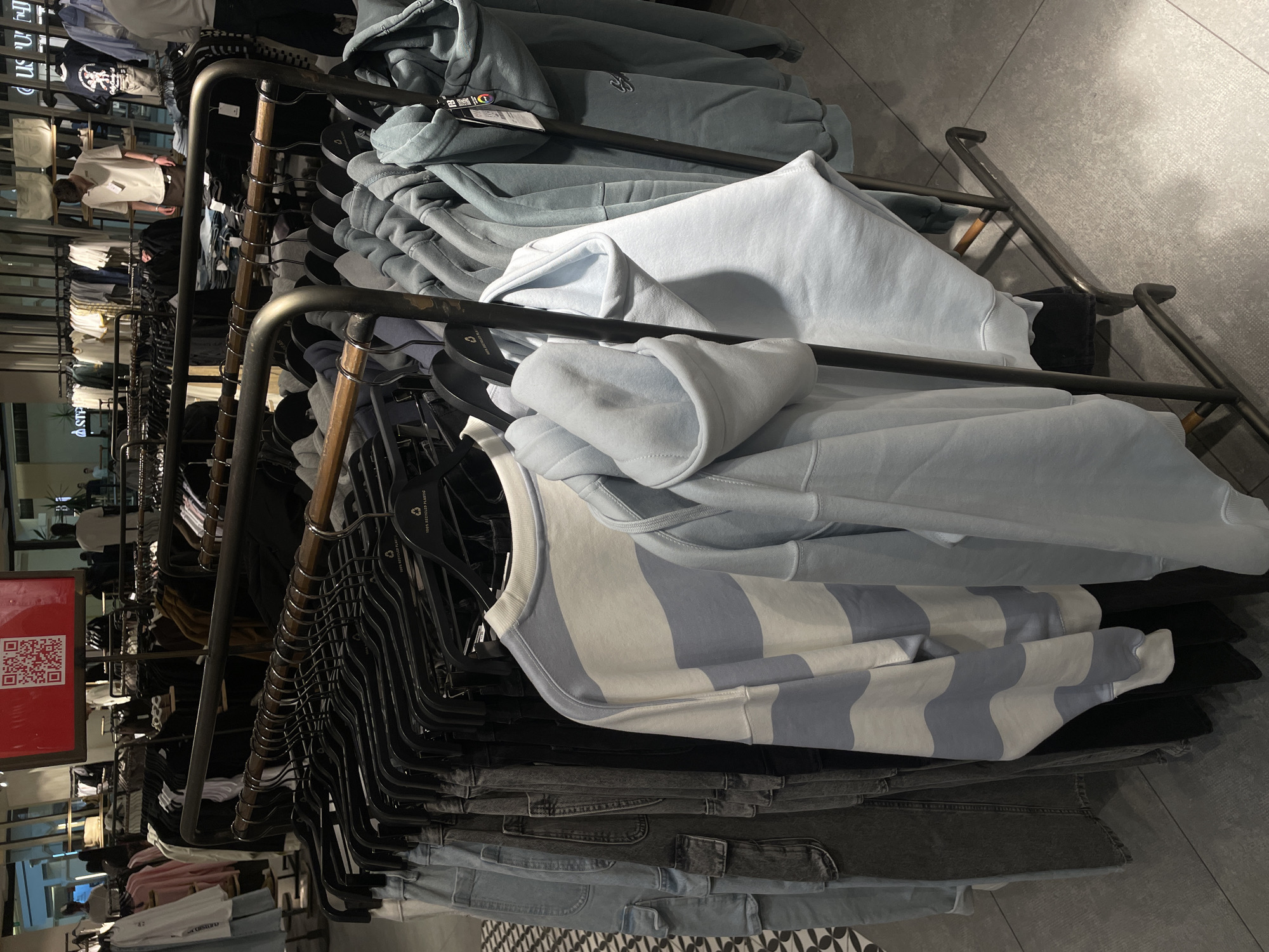 The image shows a clothing rack inside what appears to be a retail store. The rack is filled with folded hoodies and jeans, primarily in shades of blue and grey. The clothing is hung on black plastic hangers. A red sign with a QR code is visible on the left side. The location appears to be indoors, with a tile floor and other clothing racks visible in the background, possibly in Yerevan, Armenia.