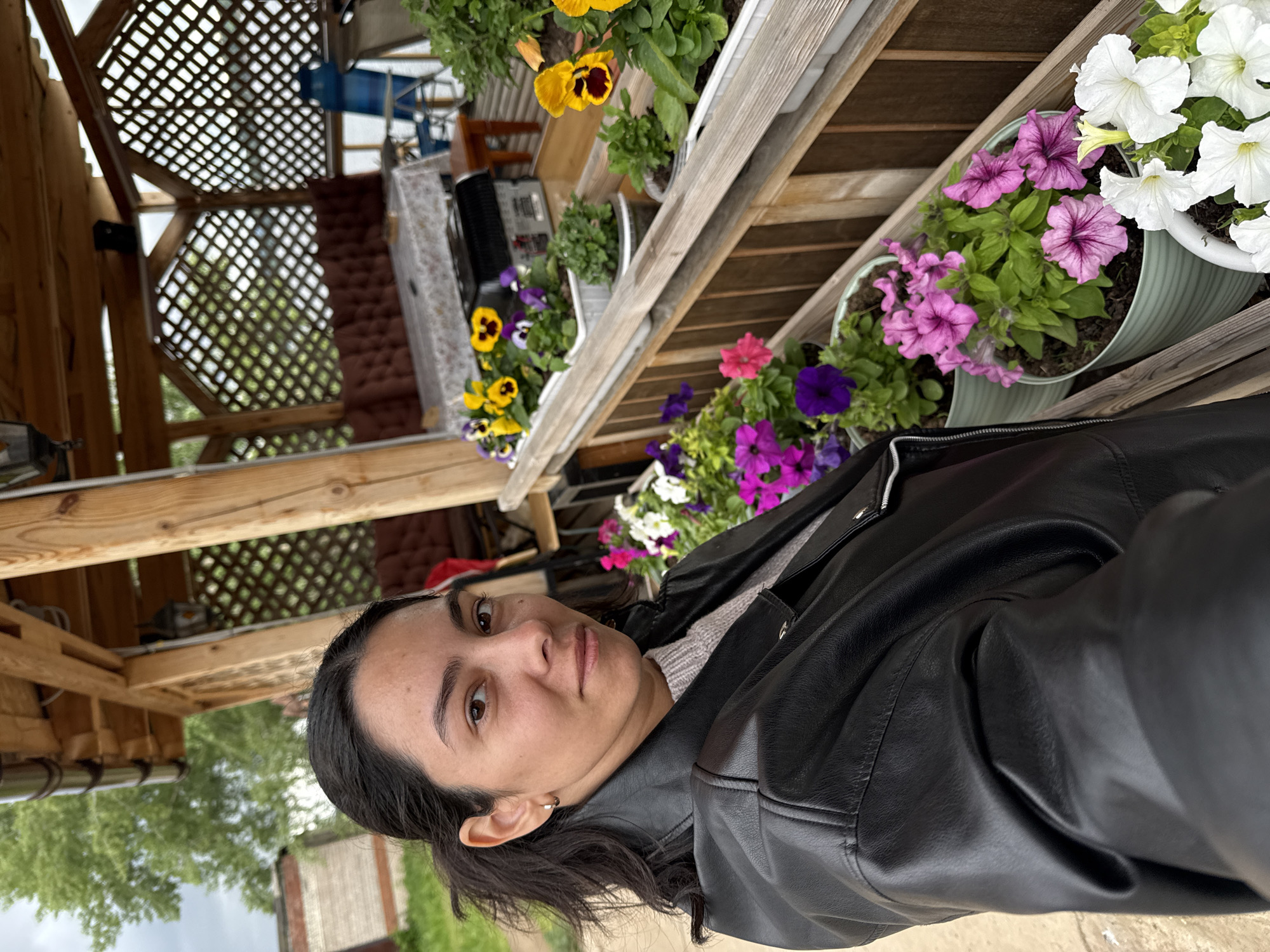 The image depicts an outdoor scene in Chaikovsky, Russia, taken from a close, selfie-style perspective. A woman with a medium-dark complexion, dark hair pulled back, and dark eyes gazes directly at the viewer with a neutral expression. She wears a black faux leather jacket over a light grey knitted top, and a small gold hoop earring is visible in her left ear. She is positioned to the left of the frame, adjacent to a light brown wooden railing.
Arranged along and behind this railing are multiple potted flowering plants. The upper tier consists of white rectangular planters containing yellow and purple pansies, along with smaller green plants. The lower tier features light green cylindrical planters holding pink and purple petunias, and a white cylindrical planter filled with white petunias.
In the mid-ground, behind the woman and the plants, a wooden gazebo-style structure is visible. This structure is characterized by exposed wooden support beams and decorative lattice panels. Inside, a long bench with dark brown, tufted cushions is situated against a wall. A table covered with a white patterned tablecloth stands in front of the bench, displaying a dark-colored appliance. Additional outdoor furniture, including a blue folding chair, is discernable further back within the gazebo. An antique-style lantern hangs from the wooden ceiling. The background, beyond the gazebo, reveals green trees, a partial view of a reddish-brown brick building, and an overcast sky.