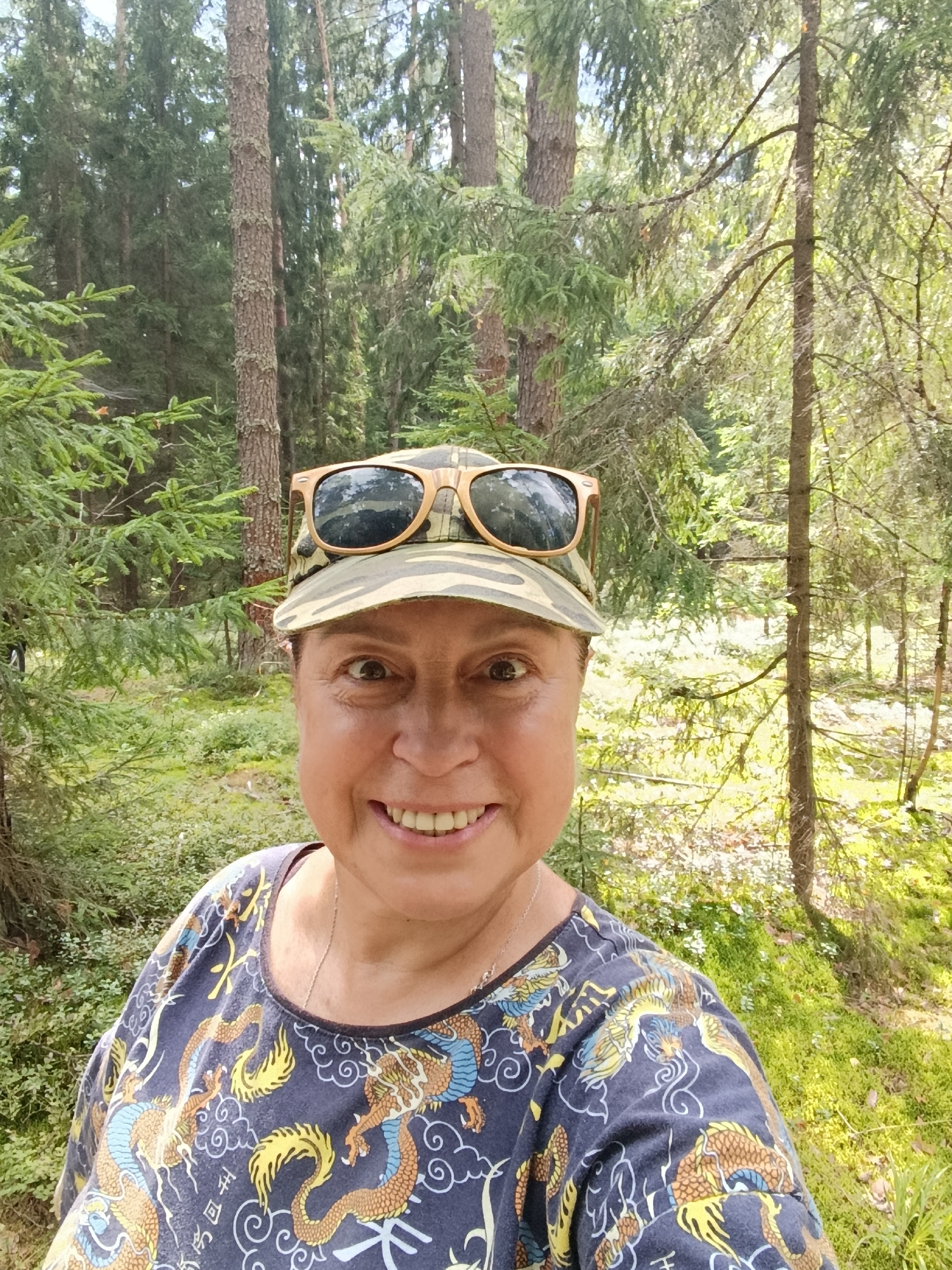 A middle-aged woman with light brown hair is smiling at the camera, taking a selfie outdoors in a forest environment. She is wearing a camouflage print baseball cap with sunglasses resting on top, and a dark blue t-shirt with a colorful dragon print. The background consists of tall pine trees and lush green undergrowth. The lighting suggests it is daytime, with sunlight filtering through the trees. The scene appears to be a casual outdoor setting, possibly a park or hiking trail, located in the city of Borduki, Russia.