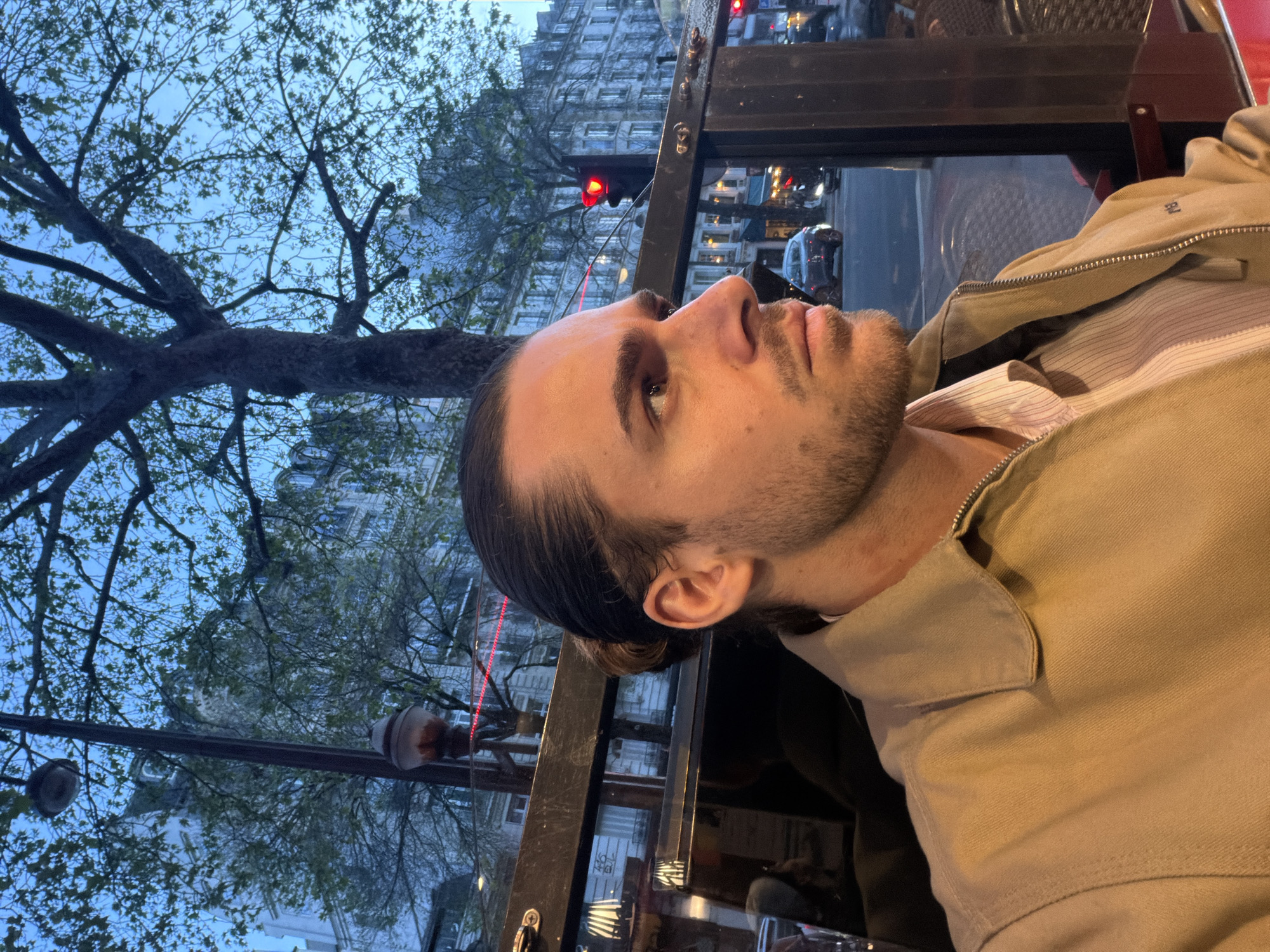 A medium shot depicts a man with long brown hair pulled back into a bun, a light beard, and light skin, viewed from the side and slightly below. He appears to be seated at an outdoor café in Paris, France. He is wearing a light-colored button-down shirt and a tan jacket. A tree with bare branches and emerging leaves is visible in the background, along with classic Parisian architecture, suggesting the setting is an urban area. A traffic light and cars hint at the activity of a city street. The shot is lit by streetlights.