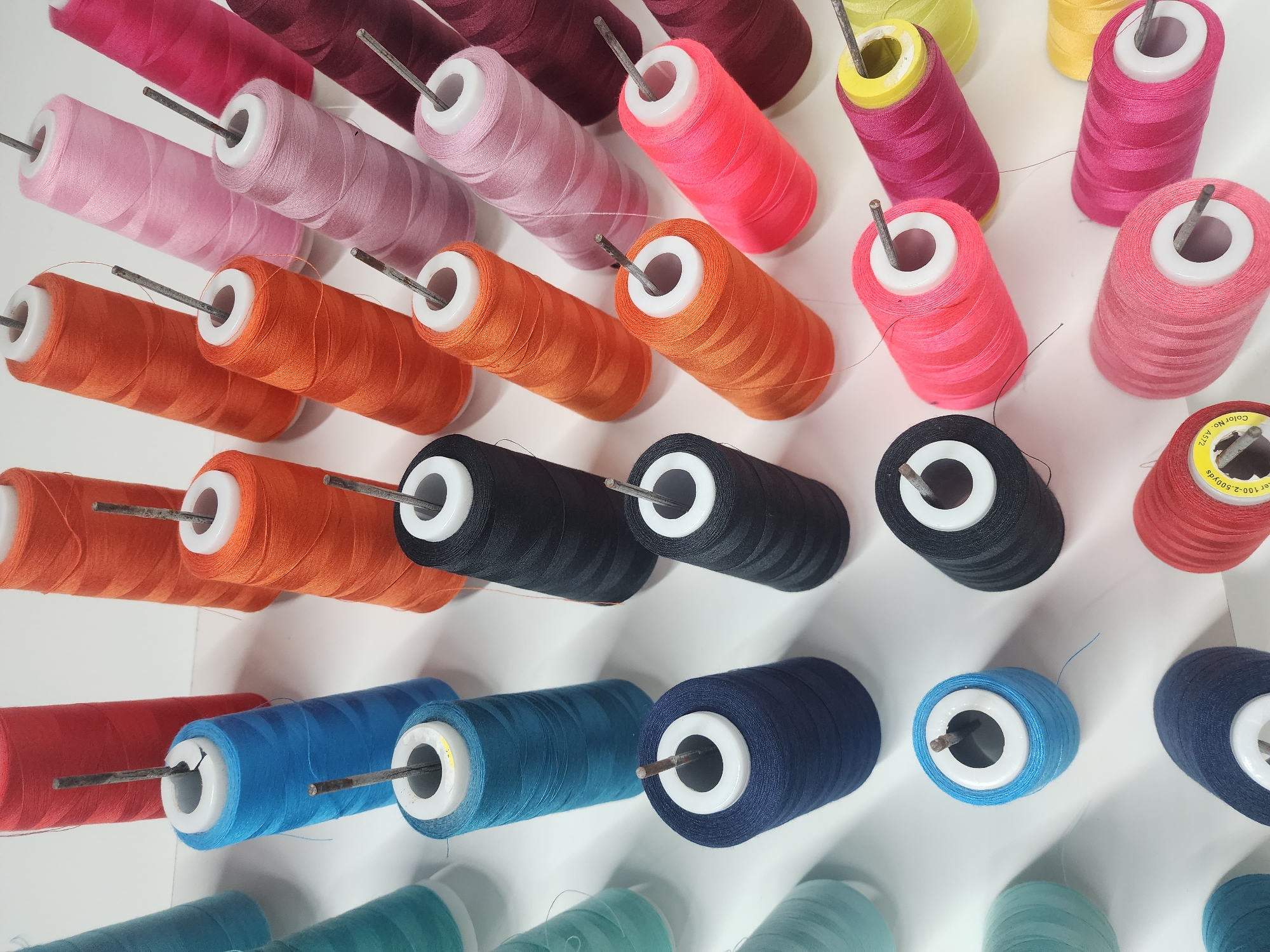 The image displays a collection of colorful thread spools arranged on a white surface. The spools are predominantly in shades of red, orange, pink, blue, teal, black, and dark blue, varying in size and winding. Each spool is pierced by a silver metal rod, with some rods angled slightly. The threads are tightly wound around the conical spools. The lighting is bright, casting subtle shadows on the white background. The scene does not provide any context or evidence to suggest a specific location within the city of Los Robles, Venezuela, or any interaction between people. It appears to be a still life arrangement of sewing supplies.