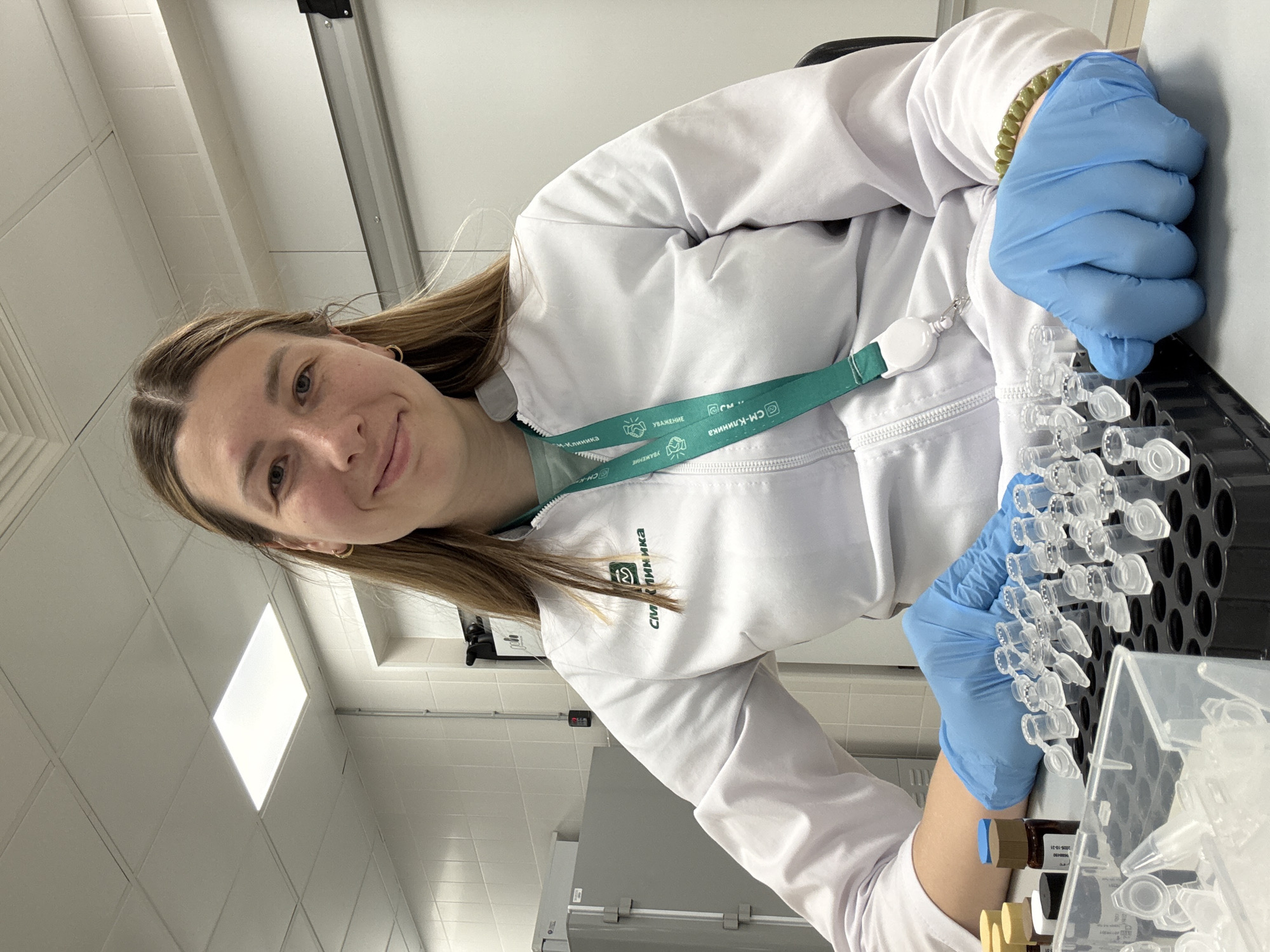 A young woman wearing a white lab coat and blue gloves is working in a laboratory. She is smiling at the camera. She is handling a rack of clear plastic tubes, possibly test tubes or vials. A green lanyard with the words "СМ-Клиника" (CM-Klinika) and "Уважение" (Uvazhenie - Respect) printed on it is around her neck. "СМ-Клиника" is a network of clinics in Moscow, Russia. The lab has white tiled walls and bright fluorescent lighting. There is laboratory equipment in the background.