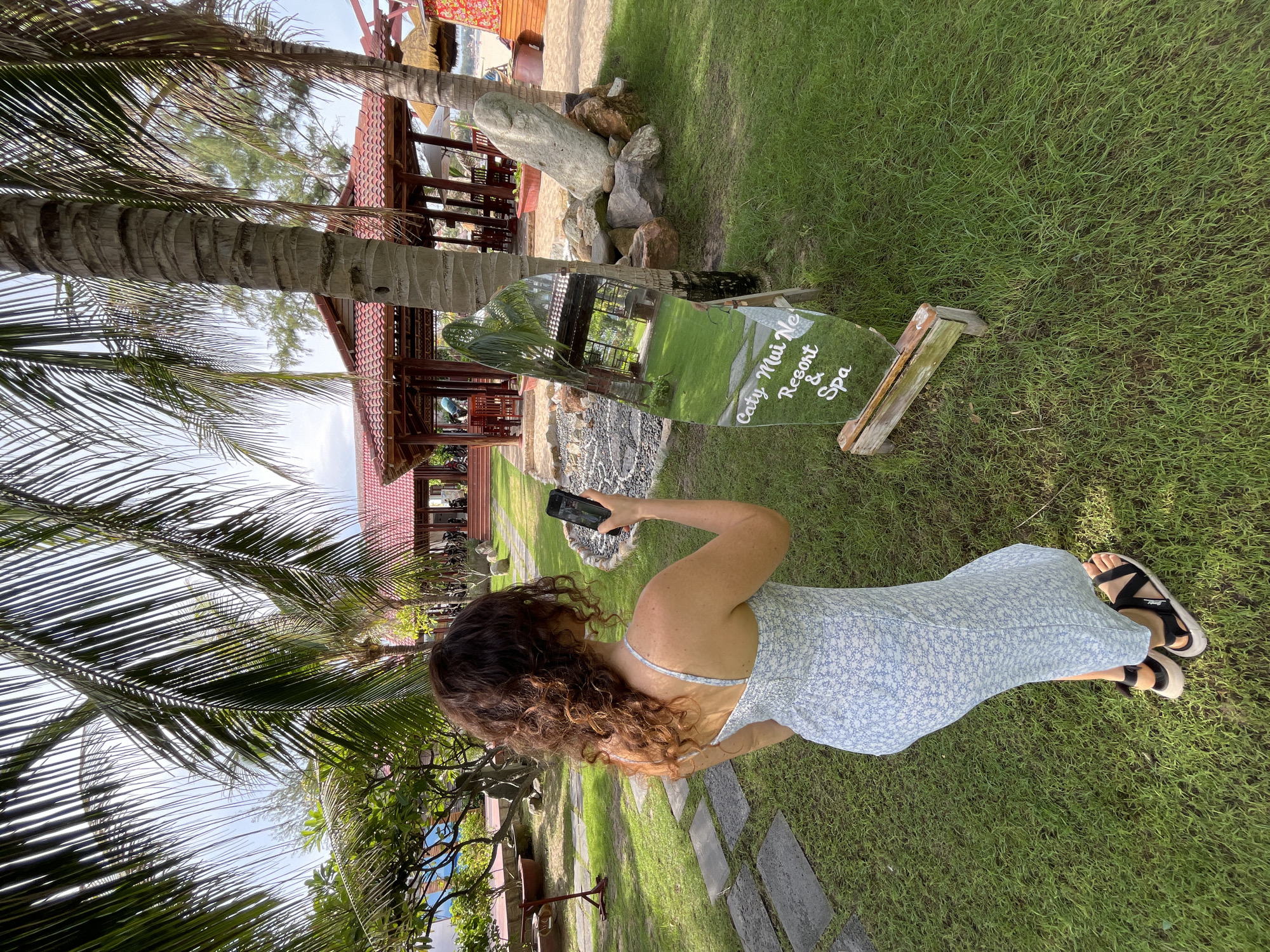 A person with curly brown hair, wearing a light blue floral dress and black sandals, is standing on a grassy lawn, holding a smartphone to take a photo of a reflective sign. The sign is shaped like a surfboard and displays white text that reads "Caty Mui Ne Resort & Spa". The sign is leaning against a wooden stand, and behind it, there are rocks and a palm tree.
In the background, there is a resort with a red tiled roof, open-air structures with wooden beams, and more palm trees. The setting appears to be tropical, with lush green grass and sand visible in the distance. The lighting suggests it is daytime, with bright sunlight casting shadows. The overall mood seems relaxed and casual, consistent with a vacation or resort setting. The location is Mũi Né, Vietnam, which is known for its beaches and resorts.