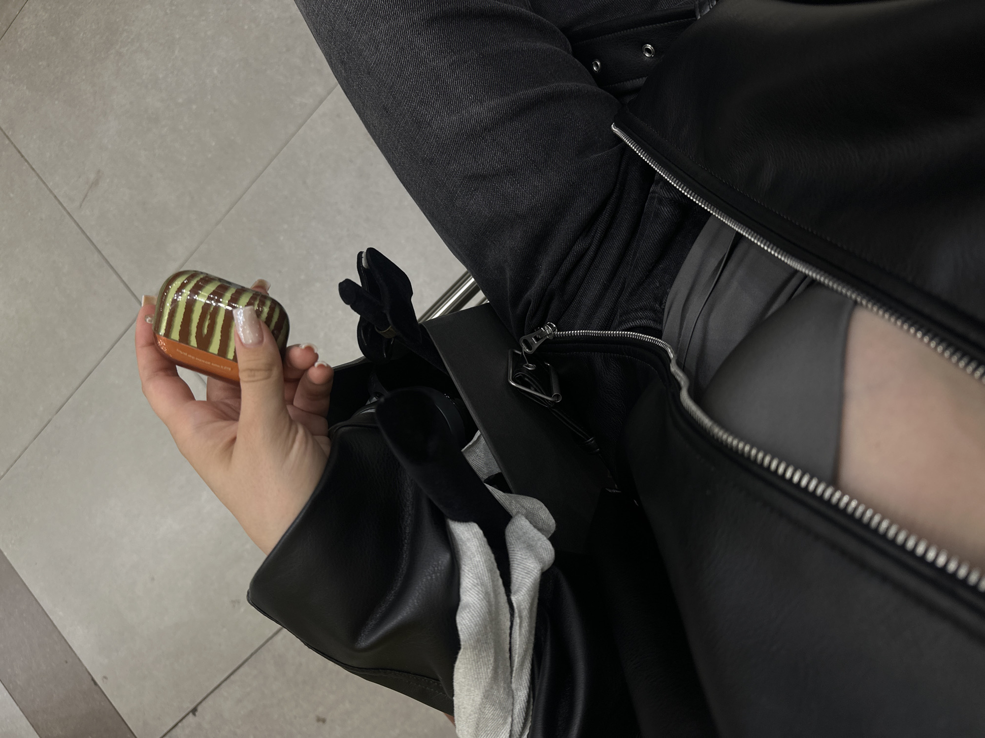 The image shows a person sitting indoors. The person is wearing dark gray jeans and a black leather jacket that is partially unzipped. A hand is holding a small, rectangular object with brown and green stripes. A black purse sits to the right of the hand, and a gray cloth is draped on the purse. The floor is light gray with a tile pattern. No specific location in Sofia, Bulgaria, can be determined from the image.