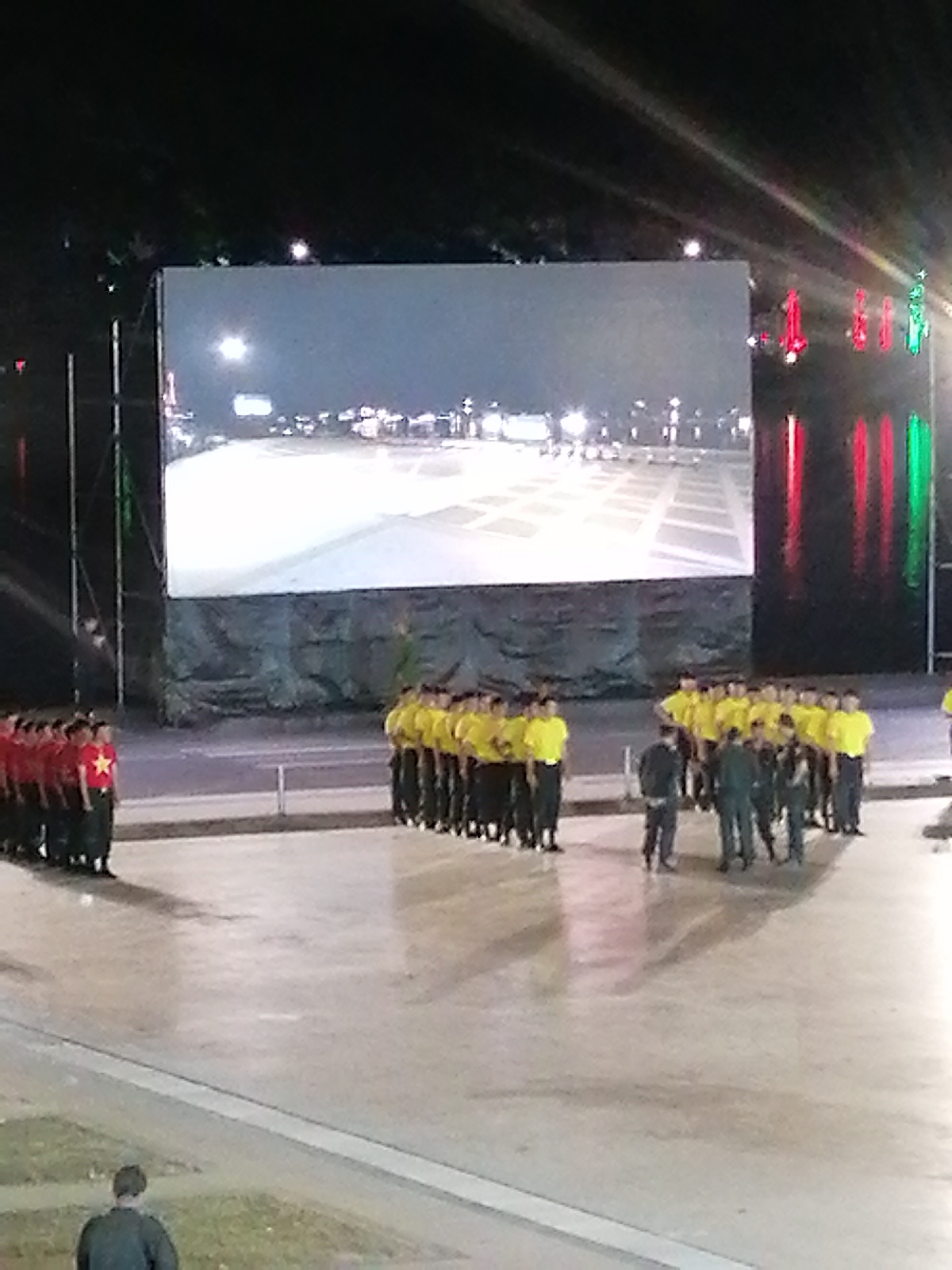 The image depicts an outdoor evening event in Da Lat, Vietnam, with a large LED screen displaying a nighttime scene of a plaza with illuminated buildings and a bright sky. In front of the screen, two lines of individuals are standing, dressed in uniforms. The line on the left is composed of people in red shirts with a yellow star, and the line on the right consists of people in yellow shirts. Several individuals in dark uniforms are positioned in front of the yellow-shirted group, and one person in a dark jacket is visible in the foreground, facing away from the camera. The ground is a paved surface. The backdrop of the scene is dark, suggesting it is nighttime, with some lights visible in the distance and on the side of the LED screen structure.