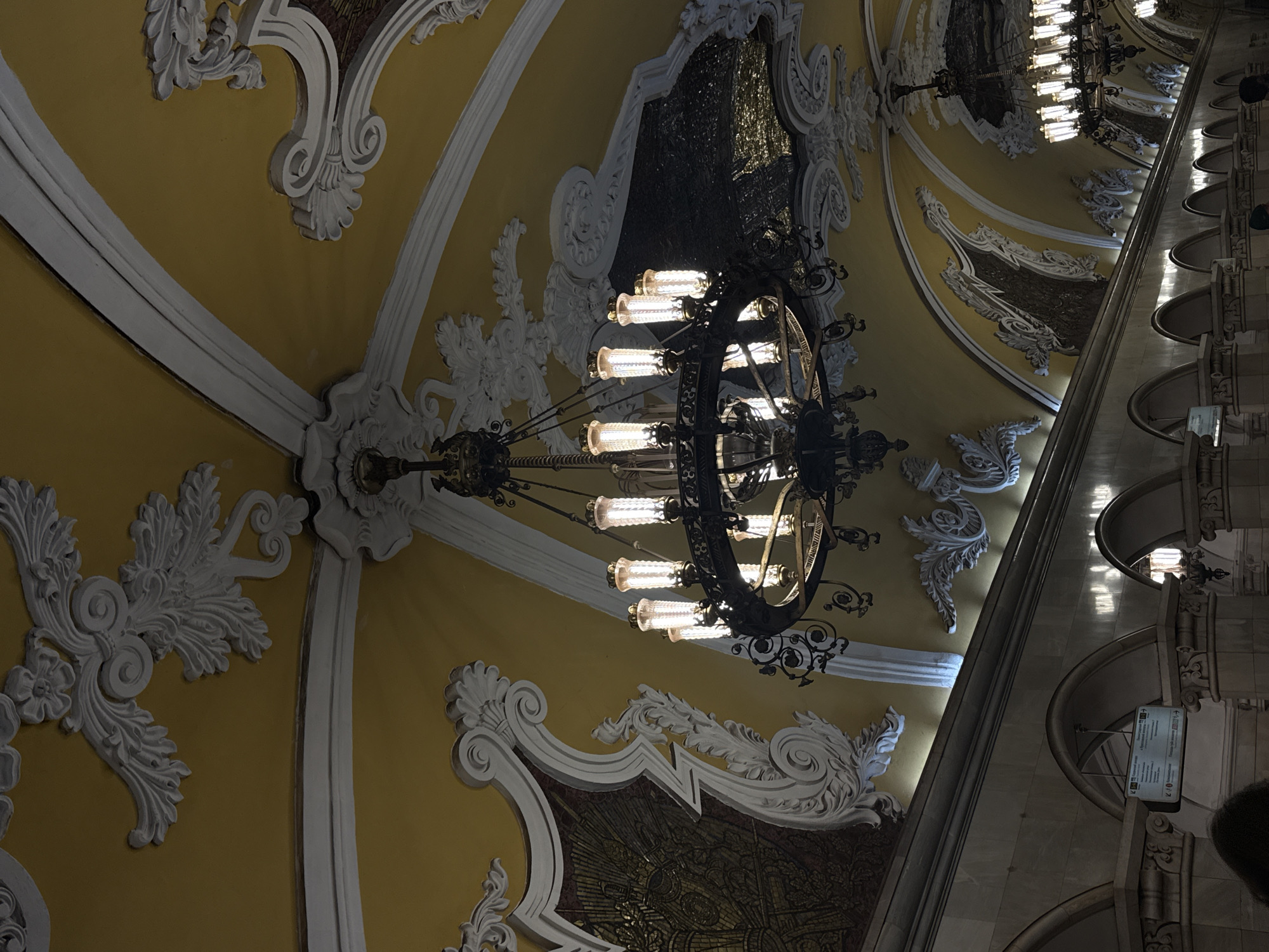 The image depicts the ornate ceiling of the Komsomolskaya-Koltsevaya metro station in Moscow, Russia. The ceiling is painted in a warm yellow hue and features elaborate white stucco moldings in baroque-inspired designs. A grand, multi-tiered chandelier hangs from the ceiling, featuring numerous cylindrical lights. A mosaic panel is visible, framed by the stucco. The station's arched structure is visible on the right side of the frame, including its pillars and informational signage.