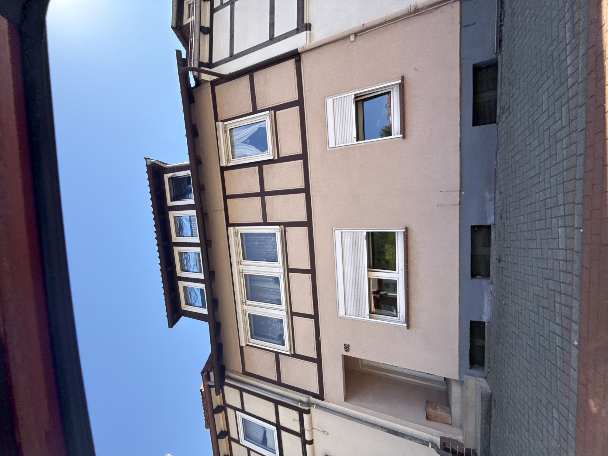 This is an exterior, eye-level, wide shot of a multi-story building in Bad Lauterberg im Harz, Germany. The building features a light pink facade with dark brown timber framing. The roofline is visible at the top, with several windows. The building has multiple windows on each level, many with white shutters, some of which are partially or fully closed. A single door is visible on the ground floor, with a small set of stairs leading up to it. The number "162" is visible next to the door. The ground in front of the building is paved with interlocking grey bricks, with a red brick border. The sky above is clear and blue. The scene appears to be a residential or commercial building, likely a row house given the adjoining structures on either side, which are partially visible and share similar architectural elements. There are no people or animals visible in the image. The environment is clean and well-maintained.