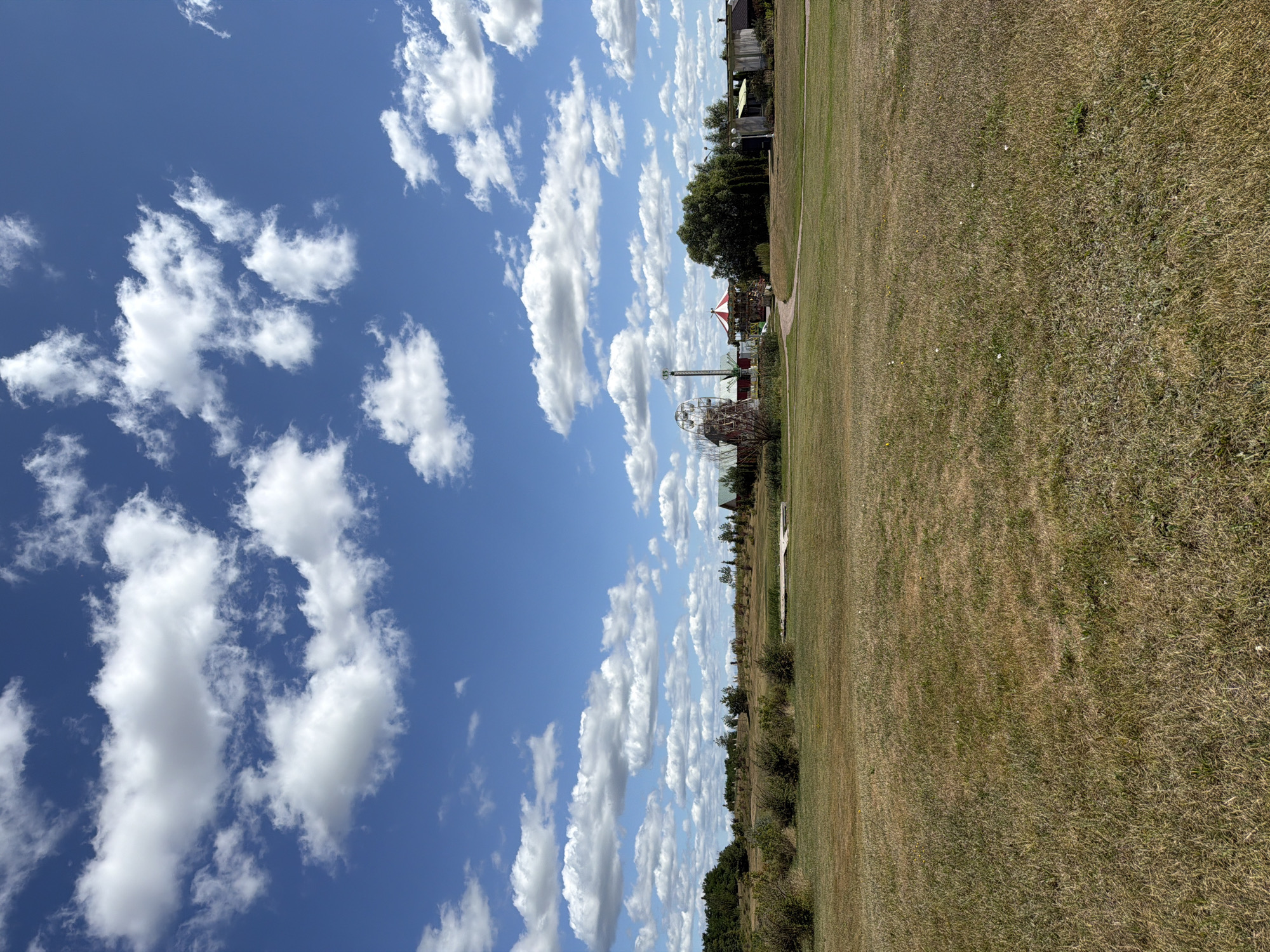 In Severynivka, Ukraine, a wide, dry grassy field stretches across the foreground under a bright blue sky dotted with fluffy white clouds. In the midground, a collection of structures suggests a small amusement area or park. Visible are a Ferris wheel, a carousel with a red and white striped roof, and several other low-lying buildings, some with reddish-brown roofs. A pathway winds through the grassy area towards these structures. A line of trees and bushes forms a border along the horizon, with more scattered greenery throughout the scene. The overall impression is of a rural landscape on a sunny day, with a touch of entertainment.