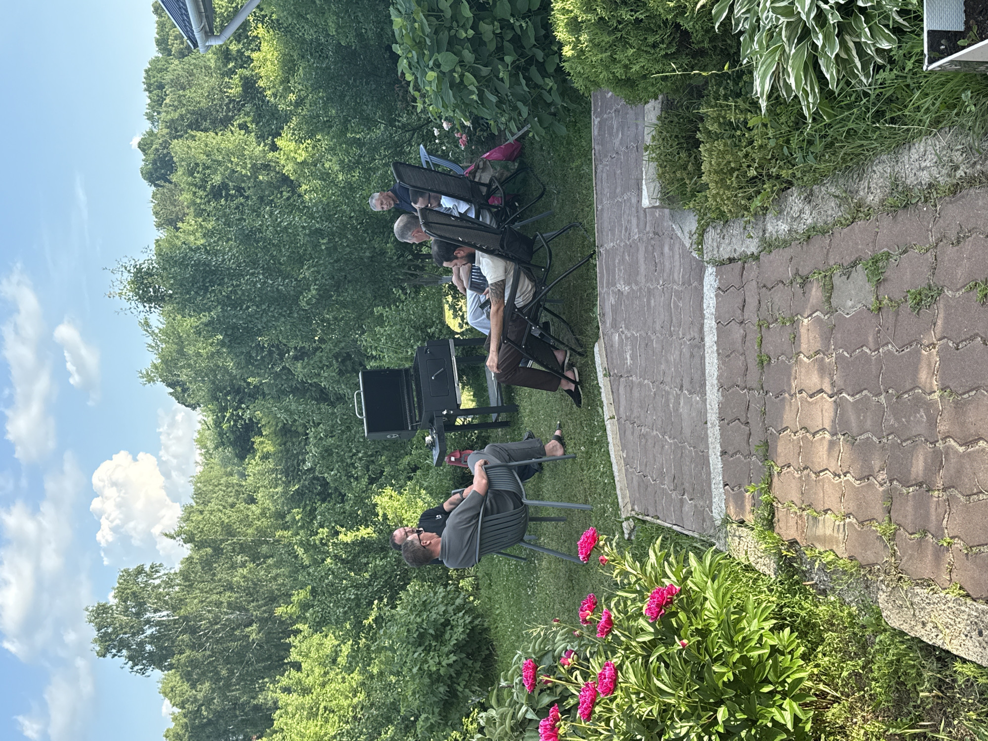 In an outdoor setting with lush green trees and foliage, a group of five men are gathered in a backyard. The men are seated in chairs, with some facing a grill in the center of the scene. The atmosphere suggests a casual social gathering, possibly a barbecue. One man is seated in a grey chair, facing left, and appears to be engaged in conversation with another man standing near the grill. Two other men are seated in black chairs, looking towards the grill. A fifth man is seated further back, also in a black chair. The ground is covered with grass, and a paved walkway made of interlocking bricks leads through the scene. In the foreground, a bush with bright pink flowers adds a splash of color to the natural environment. The sky above is blue with scattered white clouds.