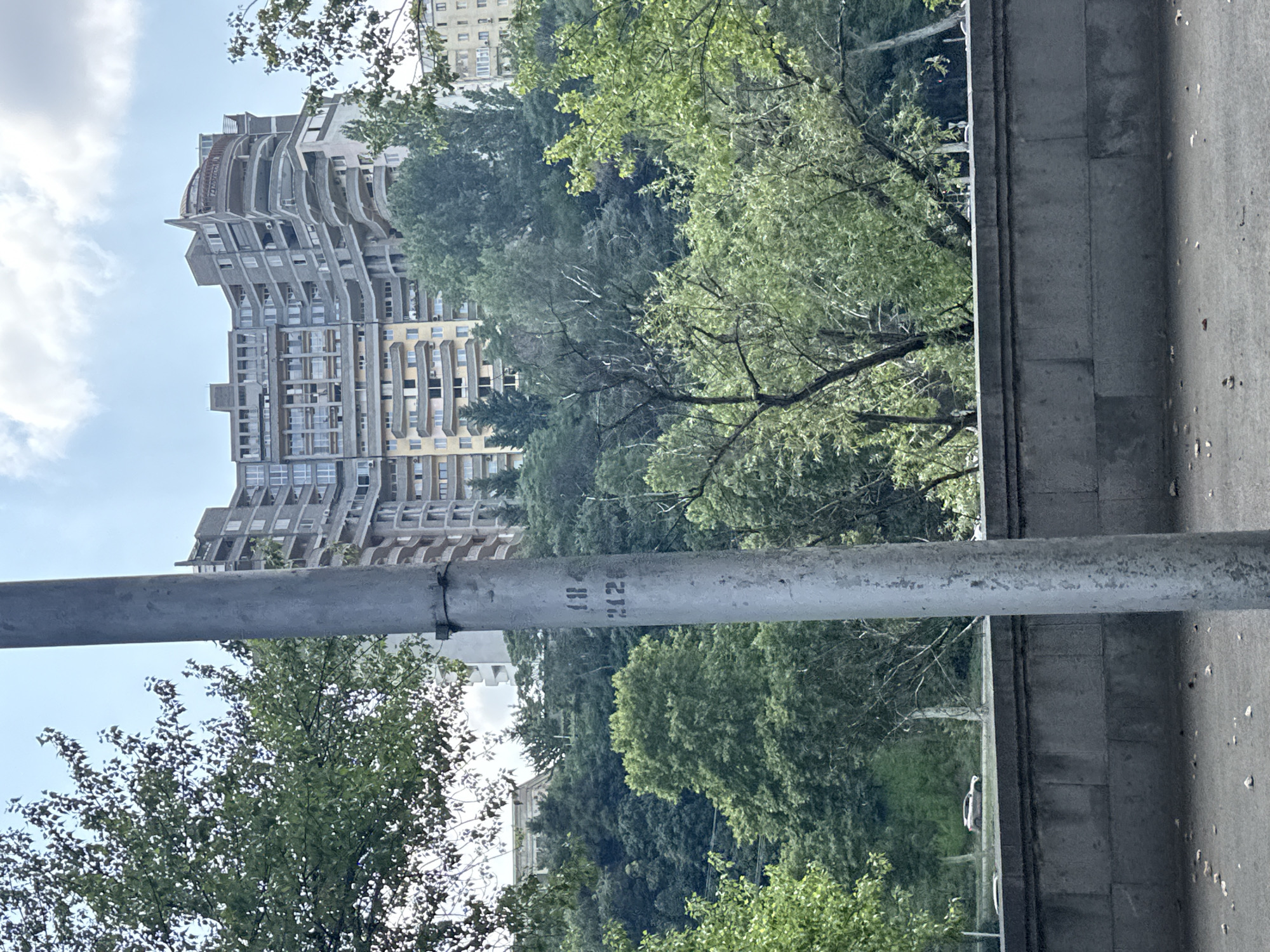 The image depicts a view of a building complex with a unique architectural style, possibly in Tbilisi, Georgia. The building is characterized by its curved facades, multiple balconies, and a stepped roofline, with a lighter-colored section dominating the lower part and a darker, more complex structure above. A grey, weathered metal pole stands in the foreground, slightly off-center to the left, with some faint markings that appear to be numbers, possibly "18" and "2120". Behind the pole and the building, a dense green hillside covered with trees is visible, with a few other buildings peeking through the foliage. A light-colored car is seen driving on a road in the mid-ground. The sky above is partially cloudy with patches of blue. The lower part of the image shows a grey, stone retaining wall and a paved ground surface with scattered leaves and small stones. The scene appears to be a daytime outdoor shot, with natural lighting.