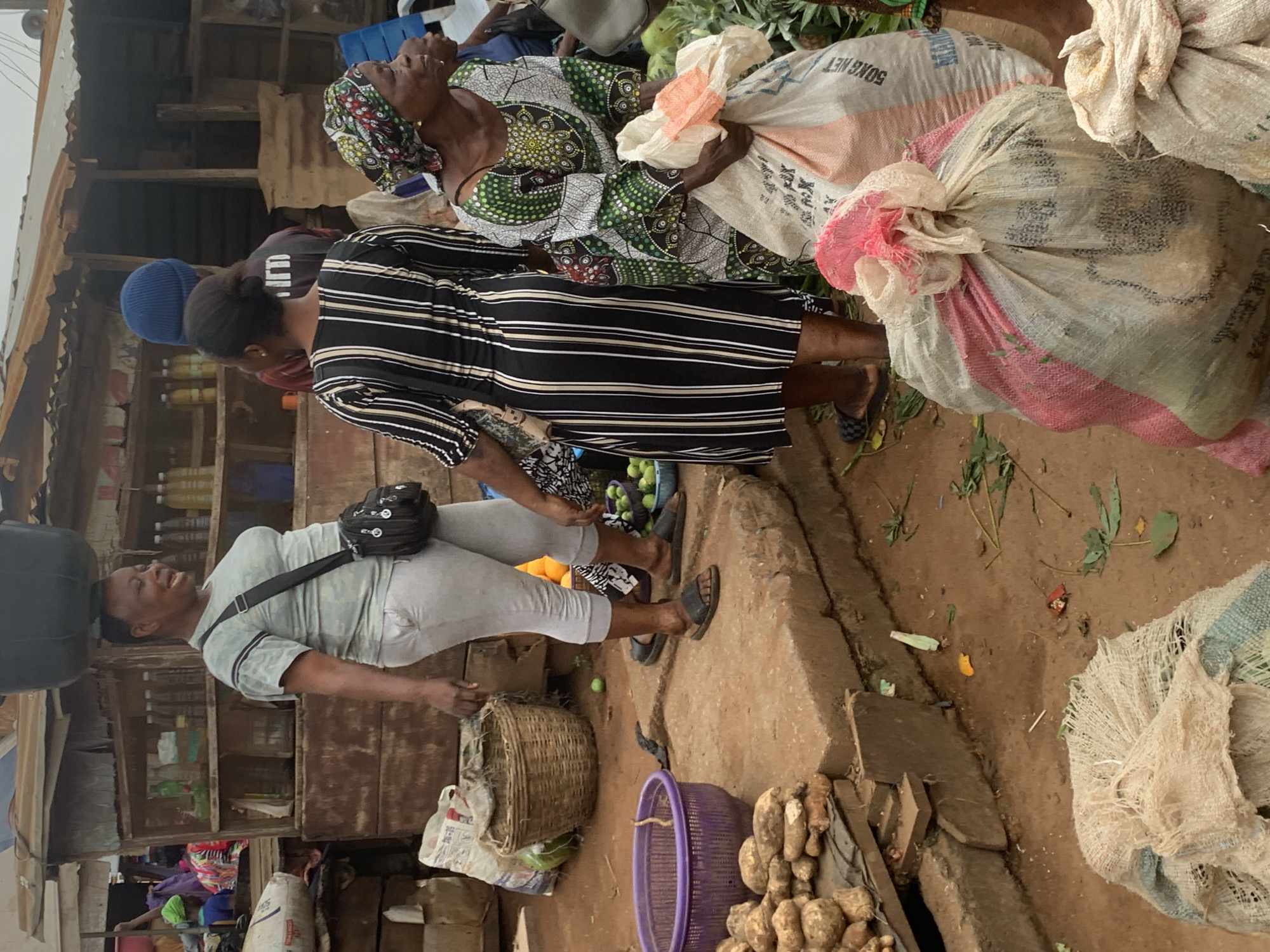 This is a medium-shot, eye-level, vertical, outdoor photograph taken in the city of Shagamu, Nigeria. The scene depicts a bustling market with several individuals engaged in commerce. In the foreground, on the right, a woman wearing a colorful headscarf and patterned dress is holding a large, off-white woven bag. To her left, a woman in a black and white striped dress stands with her back to the camera. In the midground, a woman with a large black basin on her head wears a light blue patterned shirt and grey pants, her mouth open as if speaking. Behind her, shelves in a wooden stall are stocked with bottles of liquids. The ground is dirt and uneven, with various produce, baskets, and sacks scattered around. A purple plastic basket sits on a raised concrete platform, near a pile of yams. The overall impression is of a vibrant and active marketplace.