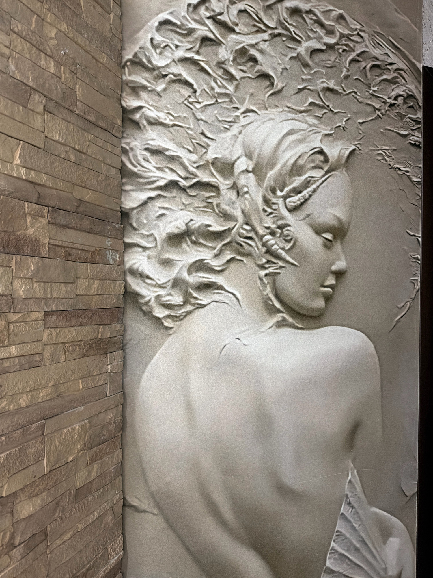 The image shows a relief sculpture of a woman's head and upper body. Her hair is styled in flowing waves, and she has delicate facial features with her eyes closed. The sculpture is monochromatic, appearing to be made of a light-colored material. It is mounted on a wall, with a textured stone finish to the left of the sculpture. The location in the city of Otradnaya, Russia, is not specified within the image itself.
