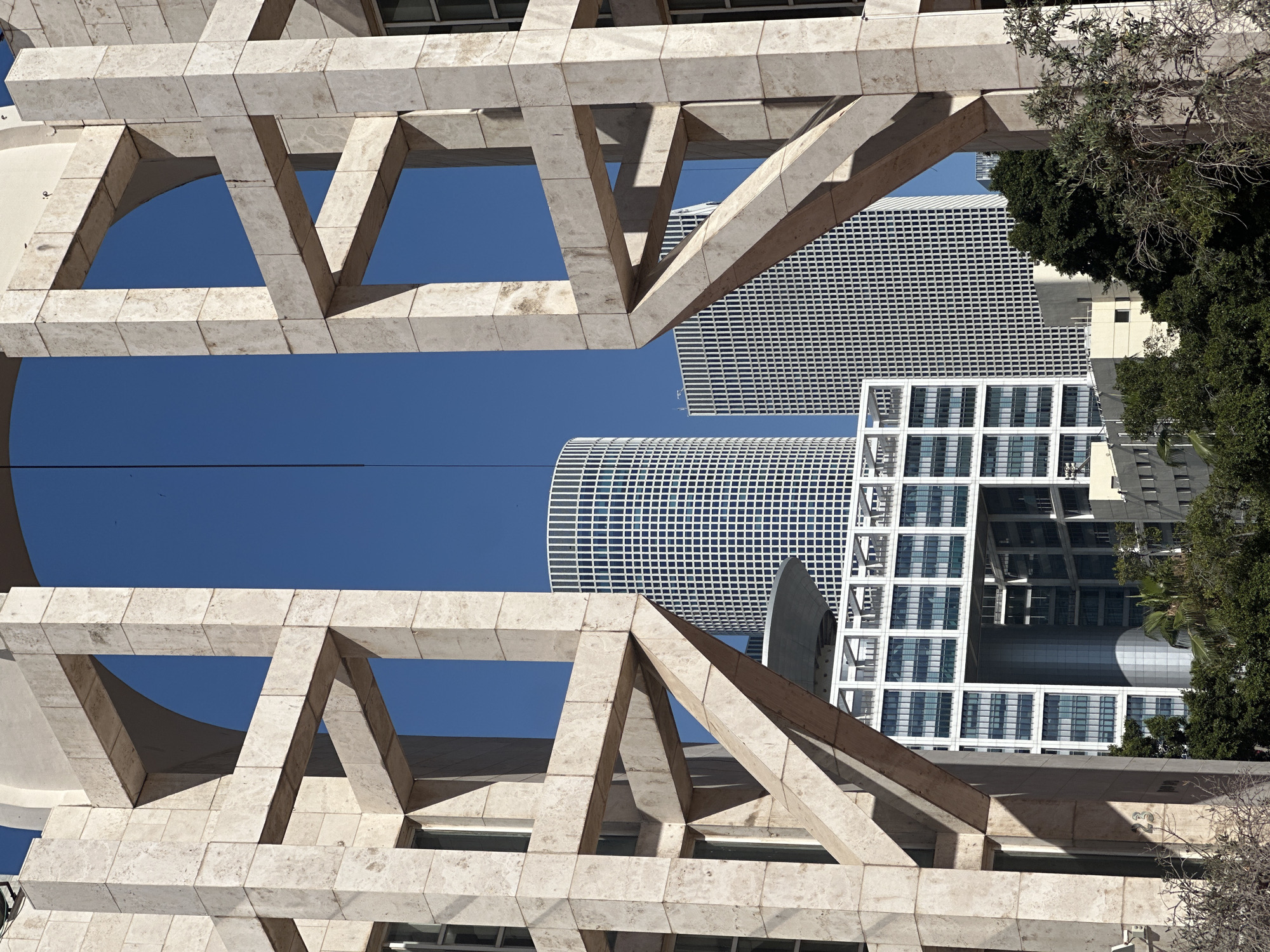 The image presents an architectural scene in Tel Aviv, Israel. The foreground features a beige-colored stone structure composed of interconnected triangular and rectangular frameworks. These structures frame a view of the sky and several modern skyscrapers in the background. The buildings exhibit grid-like facades with repetitive windows. Some trees are visible on the right side of the frame, partially obscuring one of the skyscrapers. The sky is a clear, deep blue. A thin, dark line stretches across the image.
