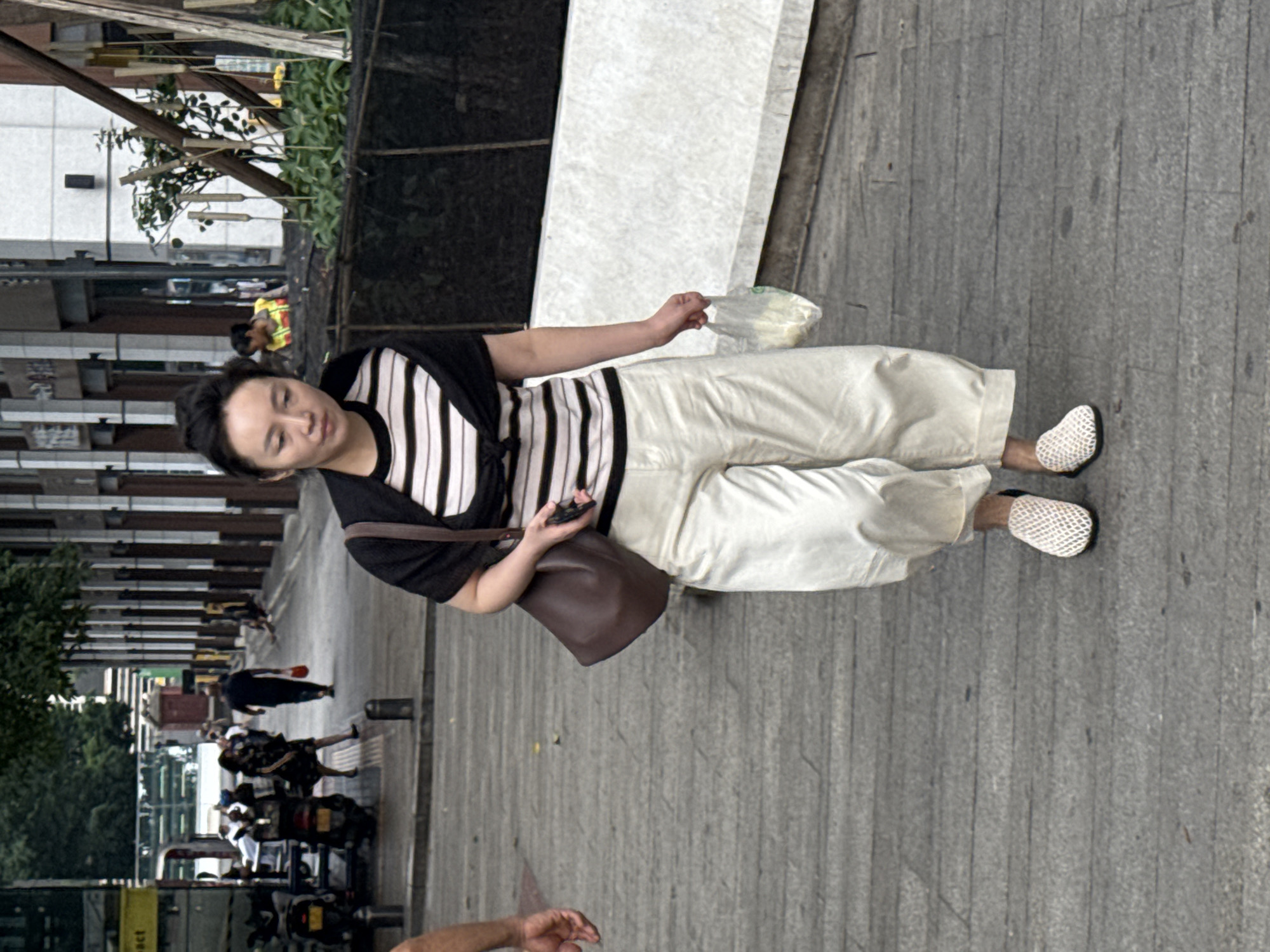 A full-body shot, oriented vertically, captures a woman walking on a paved sidewalk in Yuzhong District, China. She is dressed in a black and white striped long-sleeved shirt and loose, cream-colored pants, with a dark brown shoulder bag. Her feet are adorned with white, textured slip-on shoes. She holds a clear plastic bag in her right hand and a dark object, possibly a phone, in her left hand. Her gaze is directed downward, suggesting a moment of contemplation or focus on her belongings.
The background reveals an urban environment. To the left, buildings with horizontal architectural features are visible, interspersed with trees. The sidewalk itself is made of gray cobblestones, with some debris scattered across its surface. In the distance, other people and motorcycles can be seen, indicating a public thoroughfare. The lighting suggests it is daytime, with shadows cast on the ground, hinting at a sunny day. The overall atmosphere appears casual and everyday. No text is clearly discernible in the image.