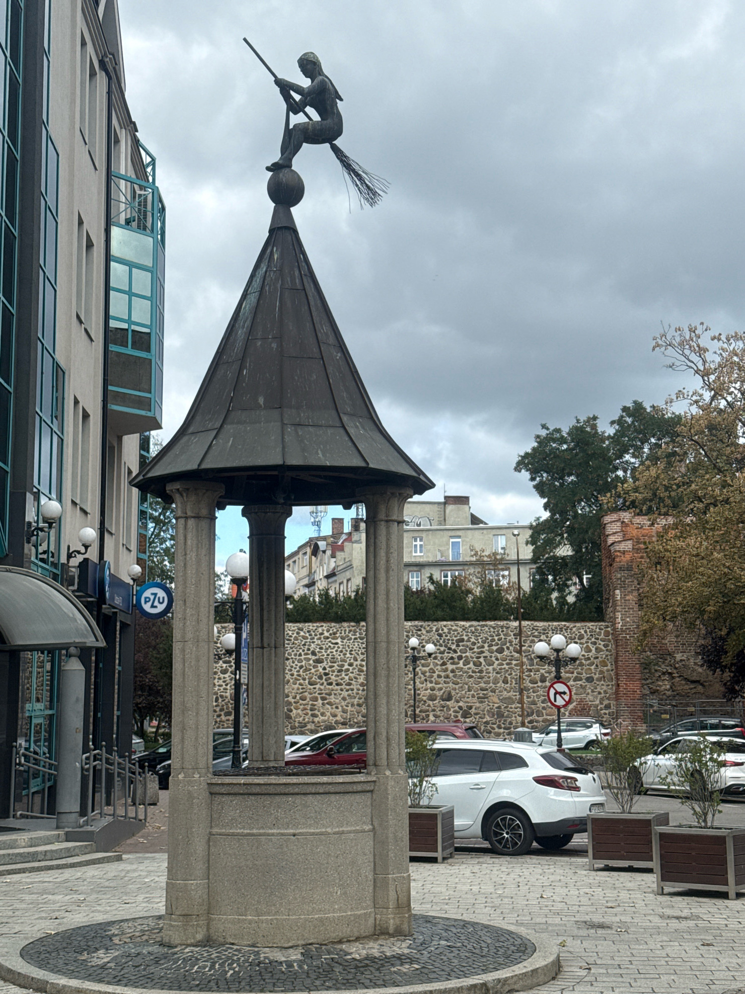 The image shows a statue of a woman on a broomstick perched atop a dark, conical structure. The statue and structure appear to be a monument or fountain in a public space. The background features a modern building with teal-tinted windows, a stone wall, trees, and parked cars. A sign with a red circle and a black arrow indicating a left turn is visible on a pole. A circular cobblestone area is in the foreground. This monument is located in the city of Gorzow, Poland, specifically in the marketplace.