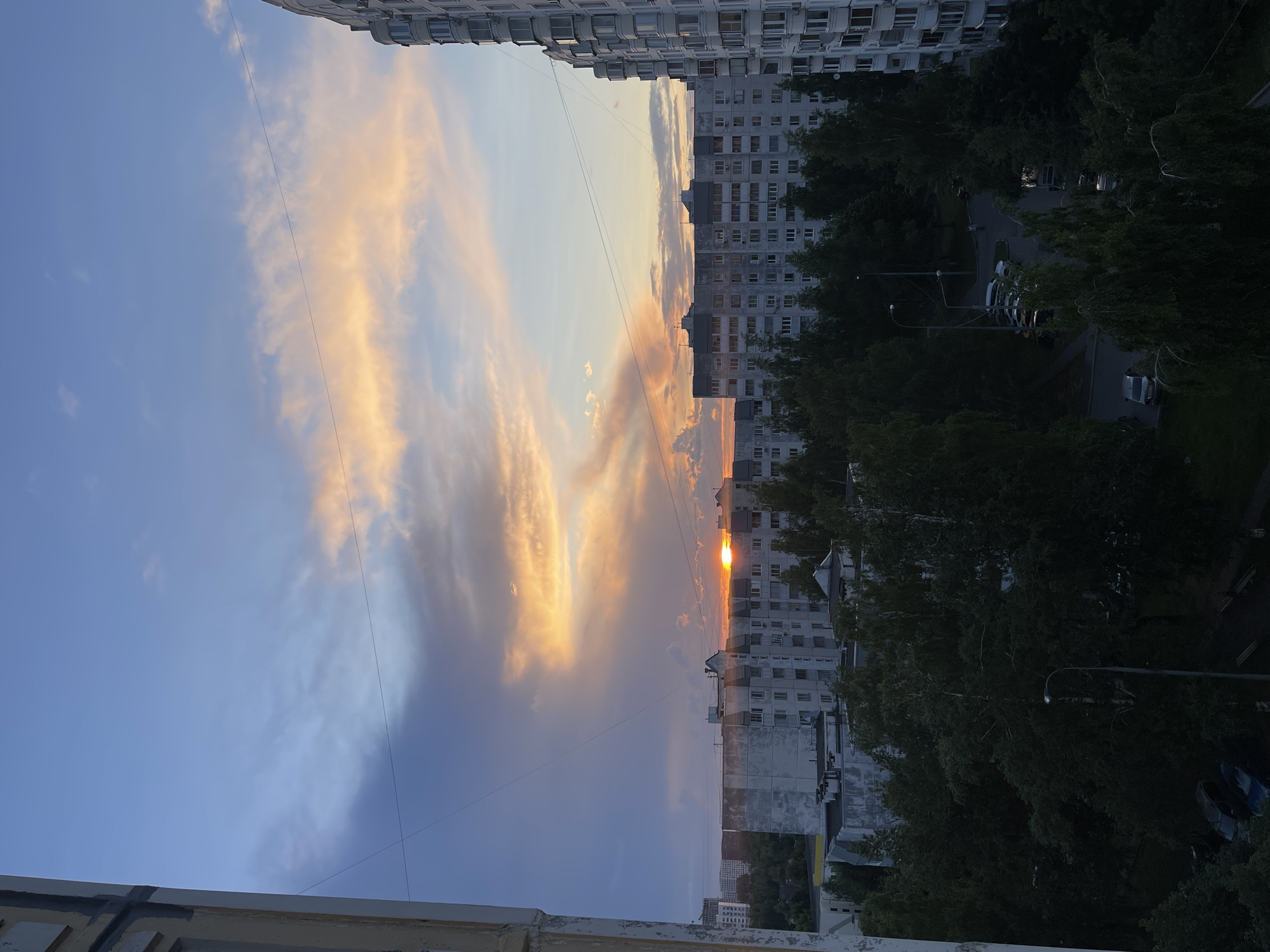 The image depicts a view of a residential area at sunset, likely in Moscow, Russia, given the architectural style of the buildings. A large, multi-story apartment building, characteristic of Soviet-era construction, dominates the right side of the frame. In the middle ground, several similar apartment buildings are visible, interspersed with trees. The sky is painted with the vibrant colors of sunset, featuring hues of orange, pink, and blue, with scattered clouds. A street lamp is visible in the foreground, along with a few parked cars and trees, suggesting a typical urban residential environment. The overall impression is a serene evening scene in a city.