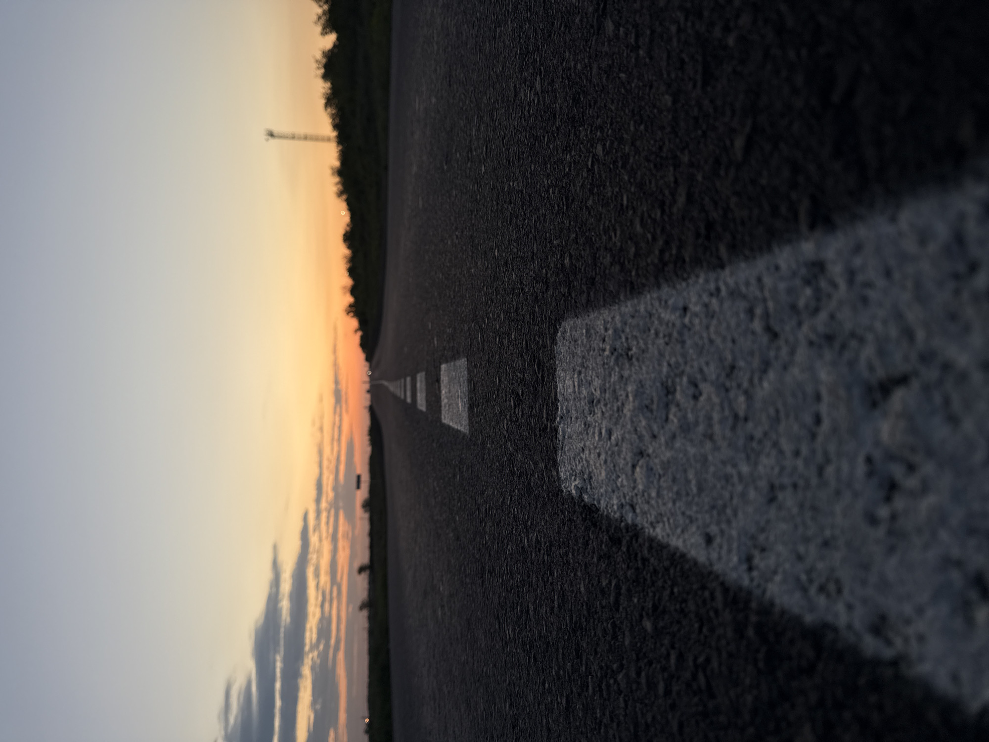 The image depicts a road stretching into the distance at dusk. The road is asphalt and has a single white lane marker on the right. The sky transitions from blue to orange. Trees line the left side of the road and the tower is in the far left.