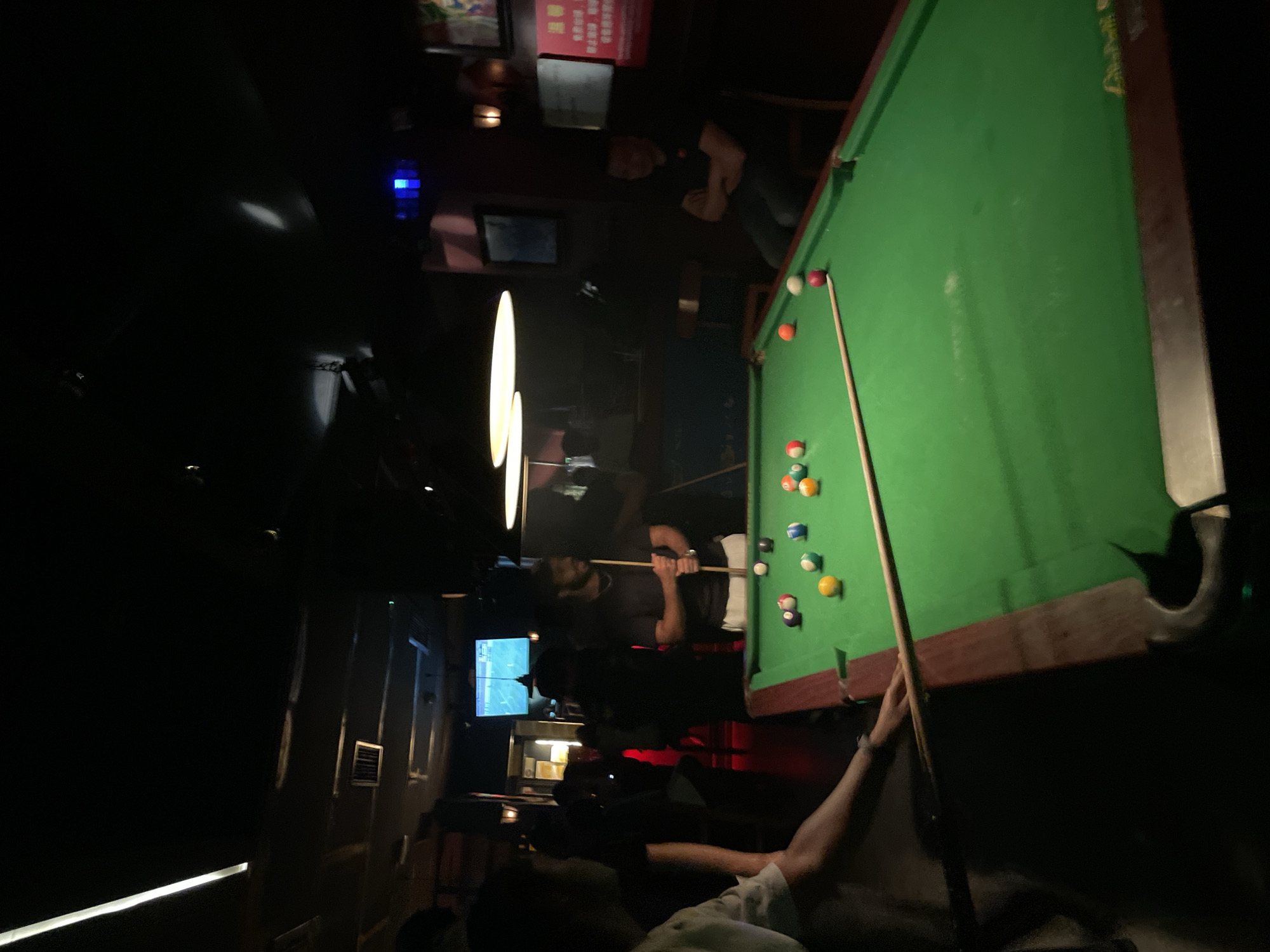 A person is playing pool on a green felt pool table. Other people are standing and sitting in a dimly lit bar in Liangjiang, China. A television is visible on the left, showing a sports game.