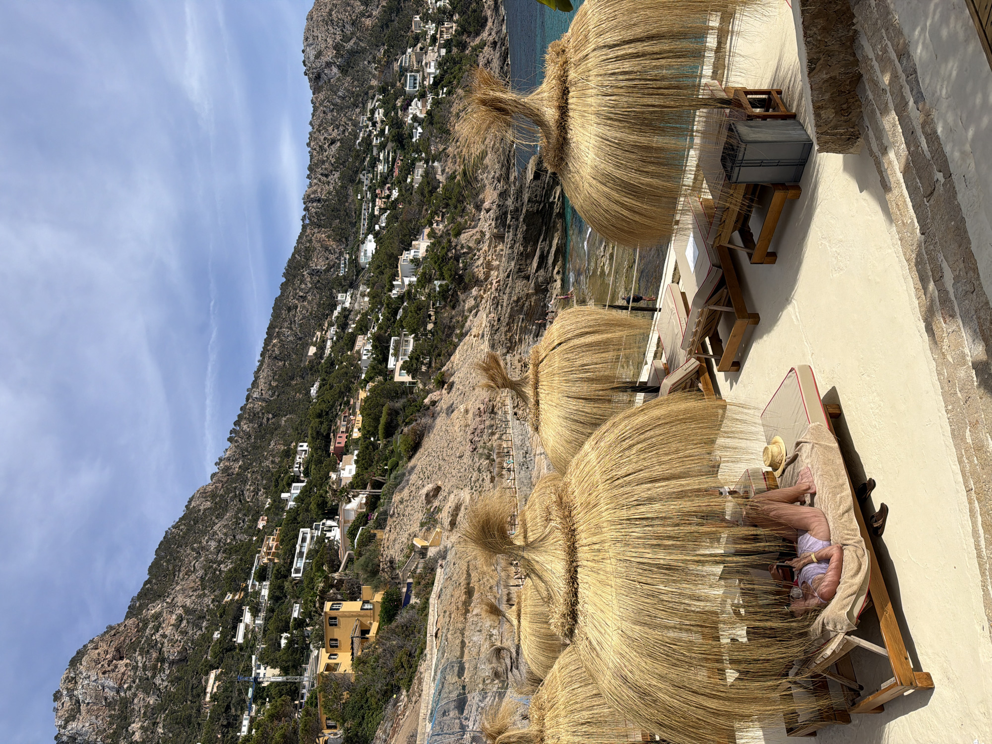 The image depicts a sun-drenched coastal scene in Port D'andratx, Spain, characterized by its hilly terrain and azure waters. In the foreground, several large, thatched umbrellas provide shade over empty lounge chairs, with one occupied by a woman in a white bikini reclining and looking at her phone. A straw hat rests on the lounge chair next to her. The beach area is paved with light-colored stone, with steps leading down to a rocky shoreline where a few people are visible. Behind the beach, the steep, verdant hillside is dotted with white and yellow buildings, characteristic of Mediterranean architecture. The sea is a vibrant turquoise, meeting the rugged coastline. The sky is partly cloudy, with fluffy white clouds against a bright blue backdrop.