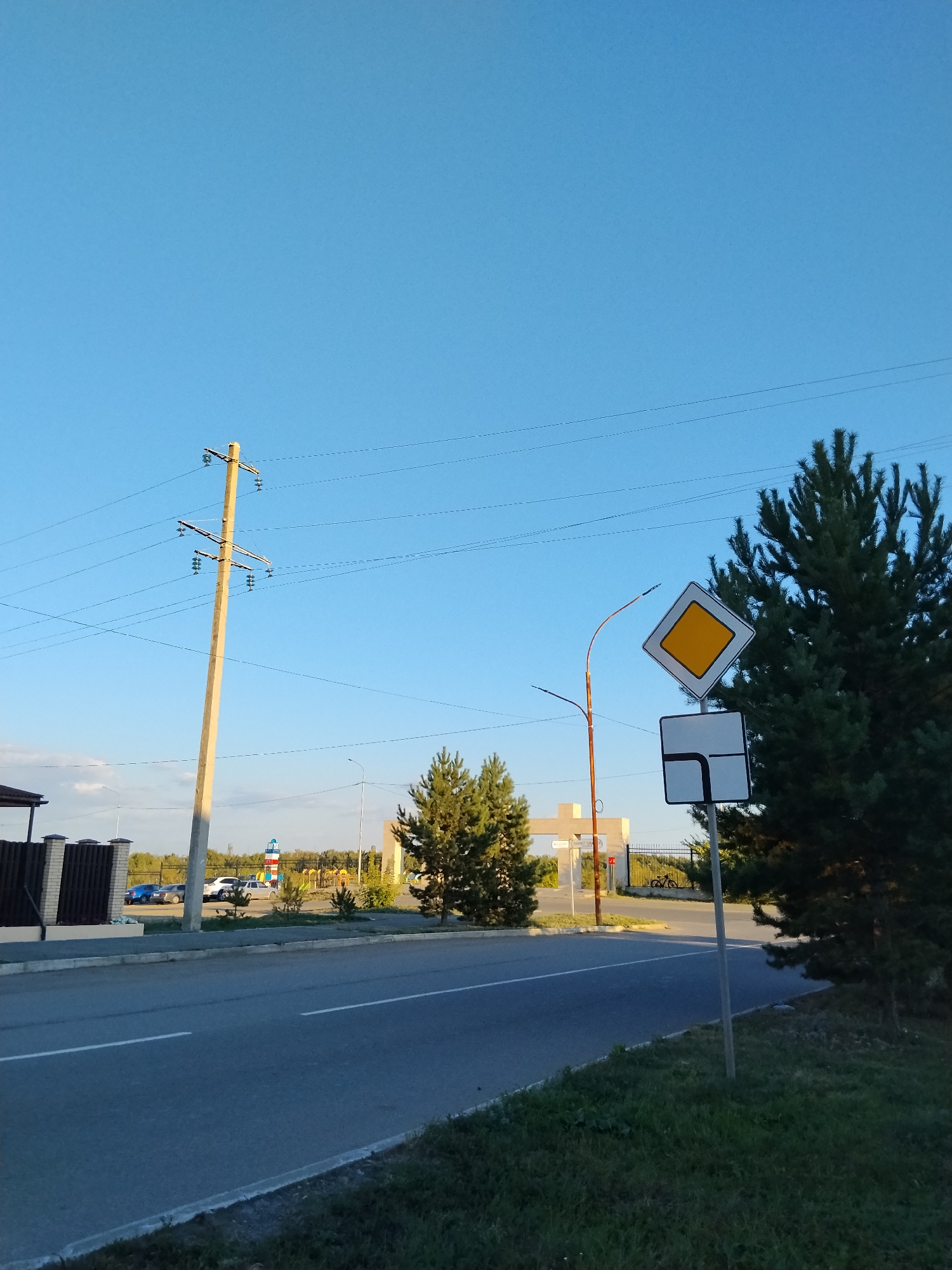 The image depicts a street scene with a clear blue sky. A concrete utility pole with electrical wires runs diagonally across the left side of the frame. In the foreground, a paved road with a white line marking the lane runs horizontally. To the right, a dense pine tree occupies a significant portion of the frame. Near the road, a traffic signpost displays two signs: a yellow diamond-shaped sign indicating a main road, and a rectangular sign with a black border and a white arrow indicating the road continues straight and turns right. Further back, a fence borders a property with cars parked nearby. In the distance, there are trees and some structures, including what appears to be a playground. This scene is located on Abylai Khan Avenue in Kokshetau, Kazakhstan, an area characterized by urban infrastructure and natural elements.