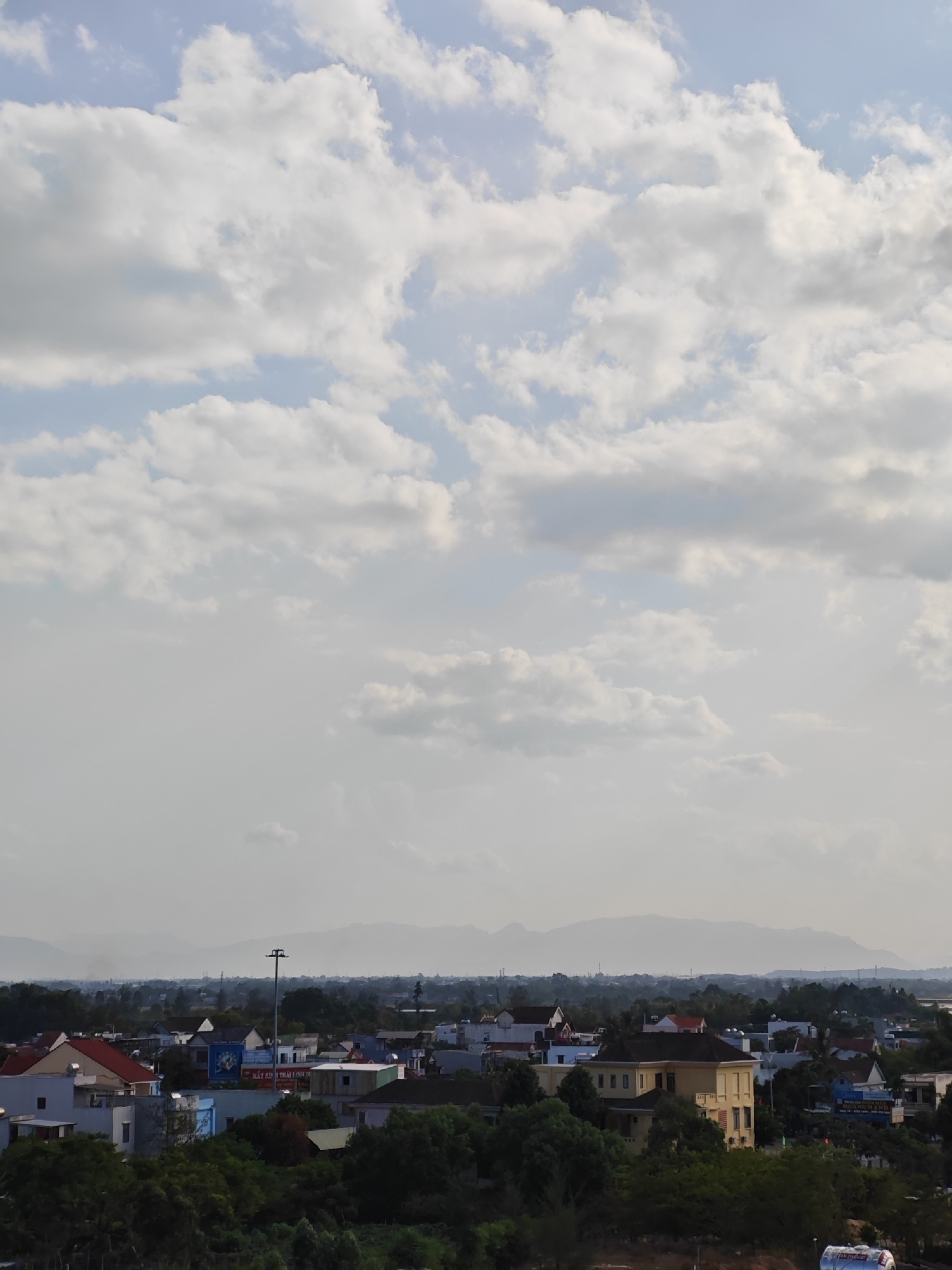 The image shows a broad view of a cityscape in Đà Nẵng, Vietnam, under a cloudy sky. In the foreground, a dense canopy of green trees partially obscures the lower portion of the buildings. The buildings are a mix of architectural styles, with some featuring red tile roofs and others with lighter-colored facades. A prominent yellow building with multiple windows stands out in the mid-ground. Several other structures of varying sizes and colors are visible, some with advertisements on their walls. In the distance, a hazy mountain range forms the horizon, and several tall poles and structures, possibly utility poles or communication towers, are scattered throughout the landscape. The overall atmosphere is serene, with the clouds suggesting a diffused, soft light. There are no people visible in the image.