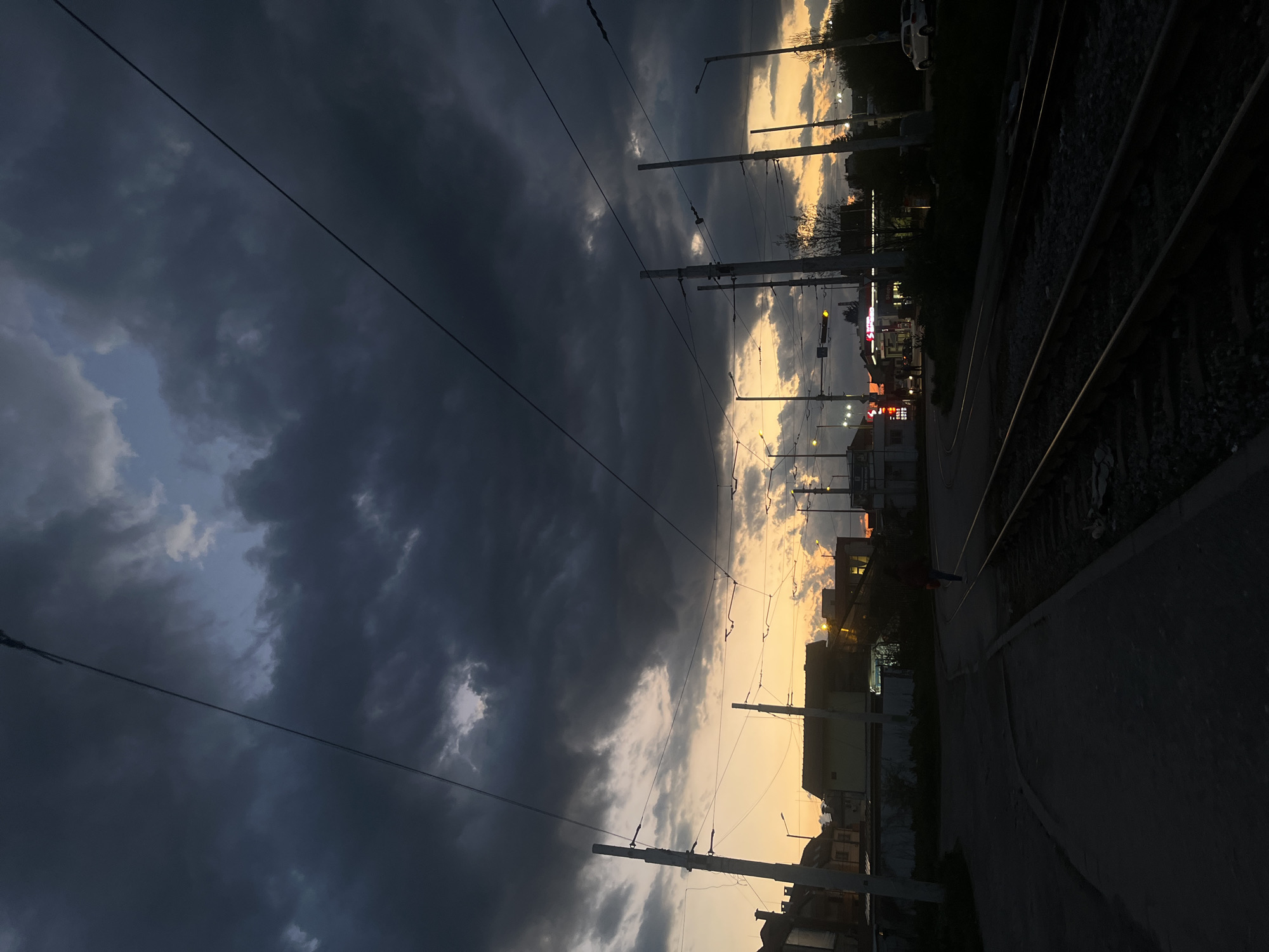 The image captures a late afternoon or early evening scene in Sofia, Bulgaria, along a tram line. Dark, heavy storm clouds dominate the left side of the frame, contrasting with the lighter, yellow-tinged sky towards the horizon, indicating a sunset or imminent dusk. The cityscape is visible in silhouette against the sky, including building outlines, streetlights, and tram overhead lines and poles. A tram track runs along the right edge of the image, with a paved area alongside it. No people are clearly visible in the image. The overall atmosphere is one of impending weather and fading light.