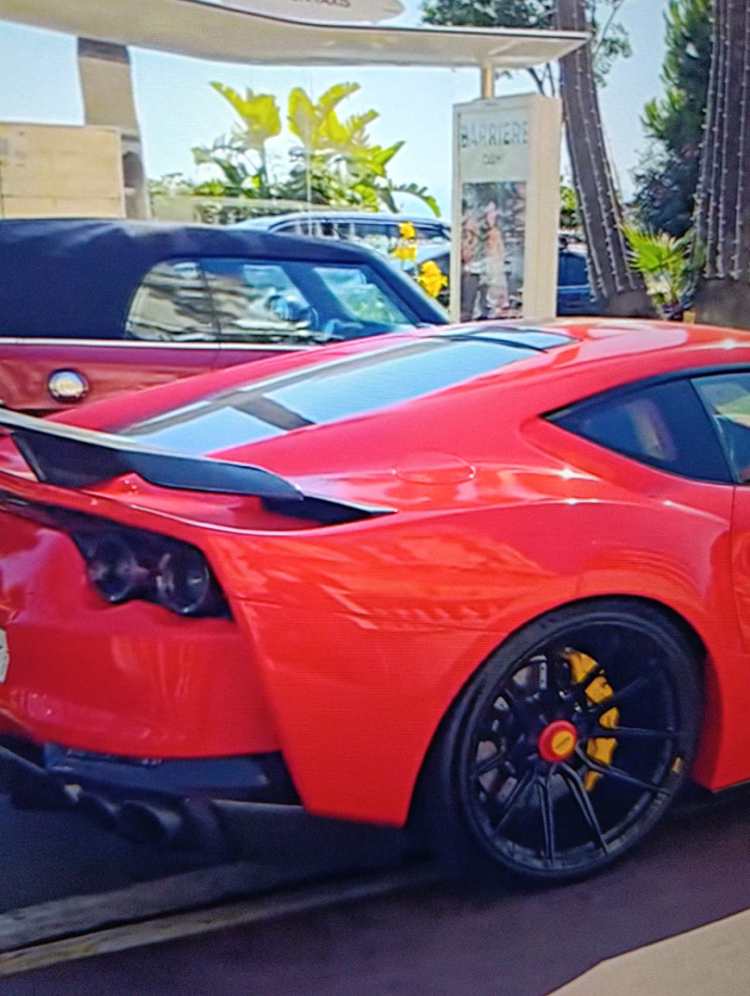 The image shows the rear of a red sports car, a Ferrari 812 Competizione, with black wheels and yellow brake calipers. The car is parked in front of a building with a sign that reads "Barriere". Other vehicles are parked nearby, including a red convertible with a dark blue top. Palm trees and other foliage are visible in the background. The location appears to be an upscale urban area. The city and region are uncertain.