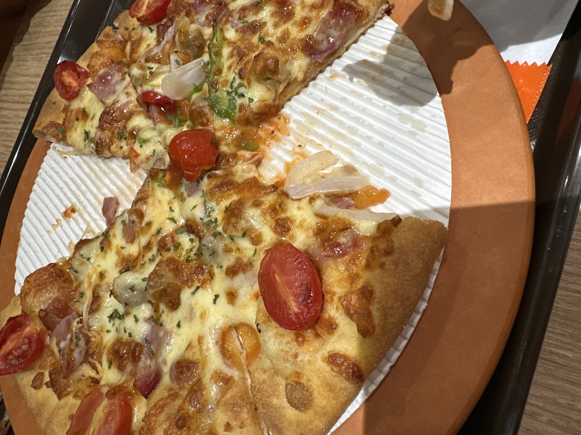 The image shows two slices of pizza on a circular, corrugated paper plate with an orange-brown edge. The pizza is topped with melted cheese, red cherry tomato halves, sliced white onion, ham, and a scattering of green herbs. The plate sits on a black tray with a wooden-patterned table surface visible in the background. The context or location in Chongqing High-tech Zone, China is not discernible from the visual information in the image. No people are visible.