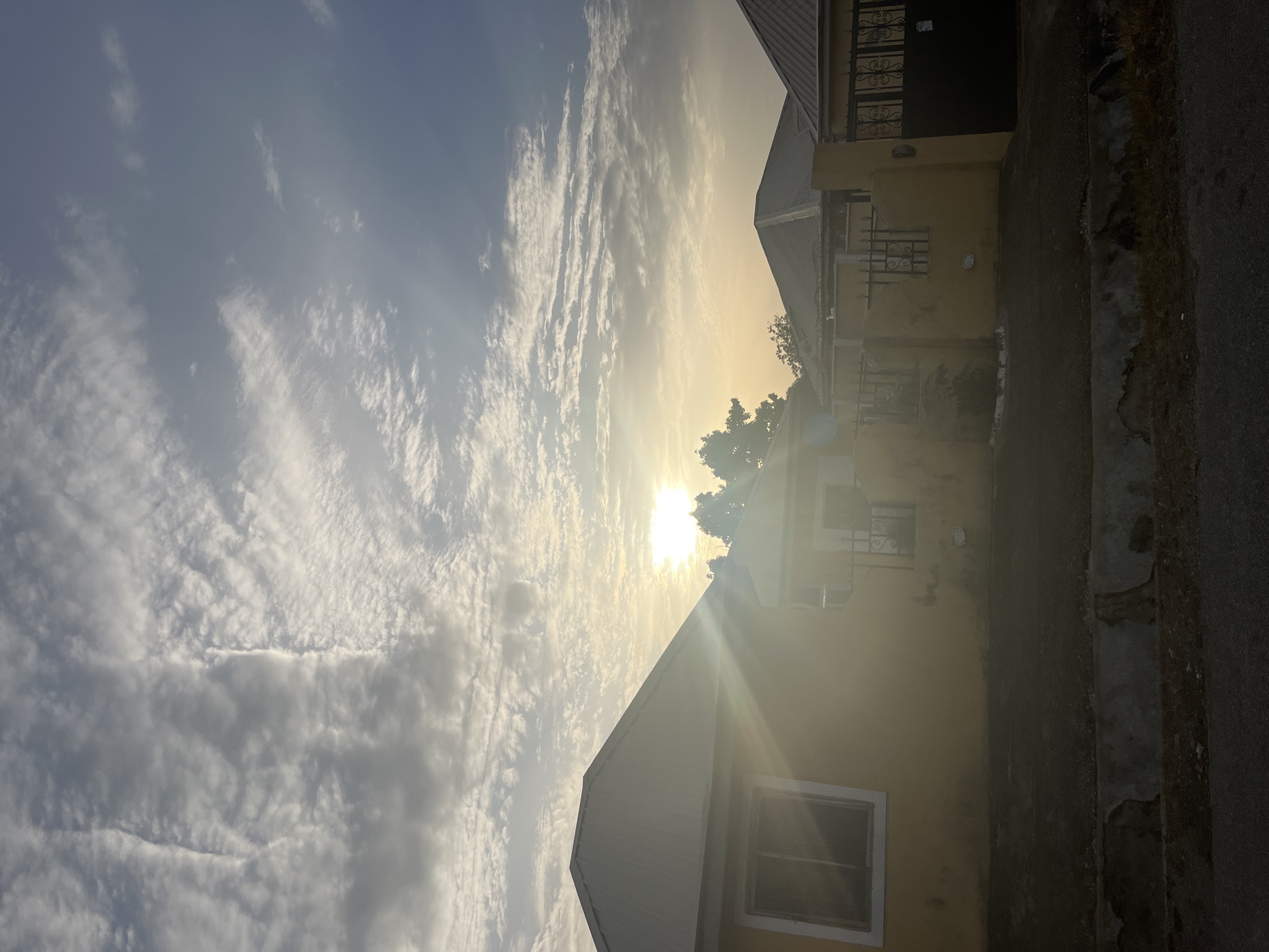 The image depicts a neighborhood scene in Abuja, Nigeria, during daylight hours. The sky is partially covered with streaky clouds, and the sun is visible, creating a bright glare. On the right side of the image are the exteriors of two buildings. The buildings are painted in shades of yellow or light brown and have windows with metal grilles. A road or sidewalk runs along the base of the buildings. The overall impression is of a quiet street scene bathed in sunlight.